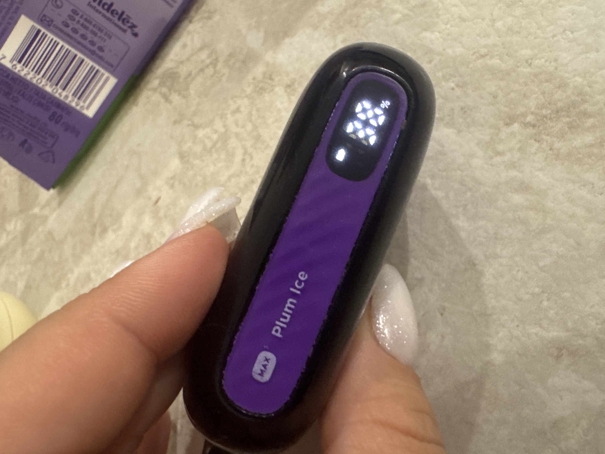 The image shows a close-up of a person's hand holding a black and purple electronic device. The device has a purple display that reads "MAX Plum Ice" and a digital readout showing "38%". The background is a textured surface, possibly a countertop. The person's fingernails are painted with white glitter polish. There is also a portion of a purple and green package with a barcode visible in the upper right corner of the image. There is no discernible information in the image that would allow for the determination of a specific location within the city of Saratov, Russia.