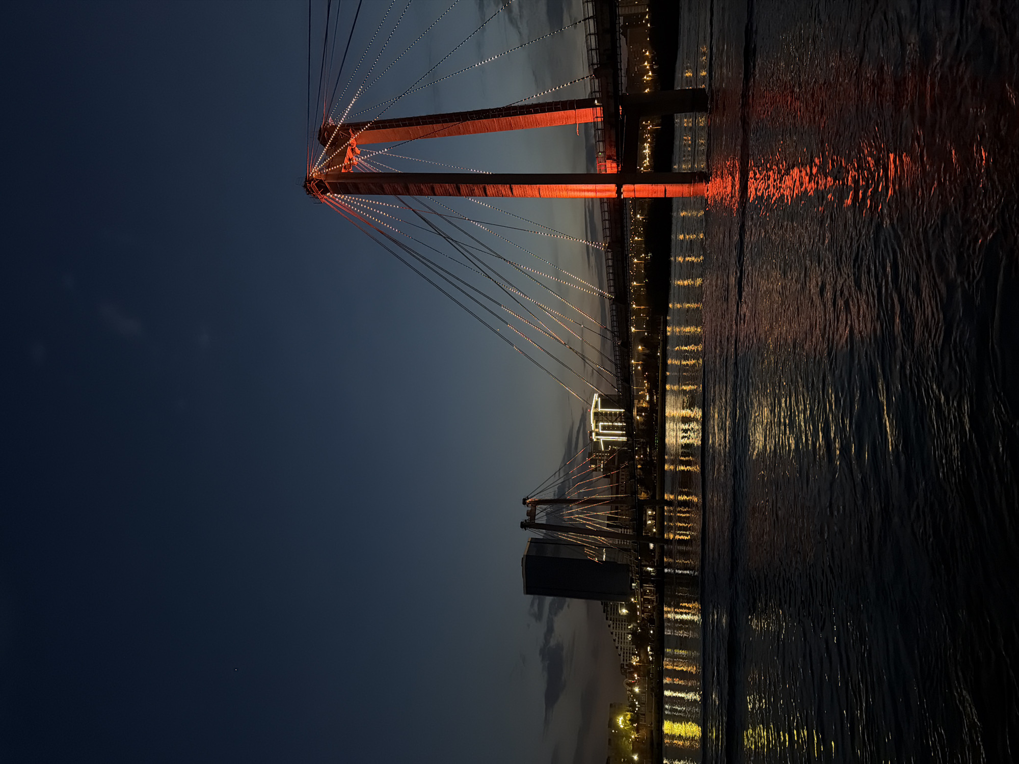The image depicts a bridge in Krasnoyarsk, Russia, at night. The bridge features a distinct red-lit tower structure with numerous cables extending from it. Reflections of the bridge and city lights are visible in the water below. A building and other lights can be seen in the background. The sky is a gradient of dark blue to lighter tones near the horizon.