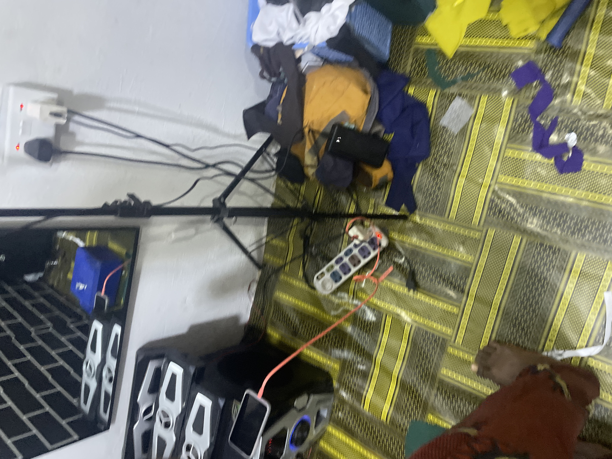 The image depicts an indoor scene, likely a room in Jalingo, Nigeria. The floor is covered with a yellow and black striped mat. In the center of the frame, a silver boombox is positioned, with a smartphone charging via a pink cable connected to a power strip. The power strip is plugged into a wall socket, which also has another appliance plugged in, both indicating red lights, suggesting they are in use. To the right of the boombox, a pile of colorful clothing, including a bright yellow garment, is scattered on the floor. A black tripod stands in the background, partially obscuring a white wall. A mirror with a brick-like pattern hangs on the wall above a blue electrical box. A person's legs and feet, wearing dark red patterned clothing, are visible in the lower left corner. The overall impression is of a somewhat cluttered living space.