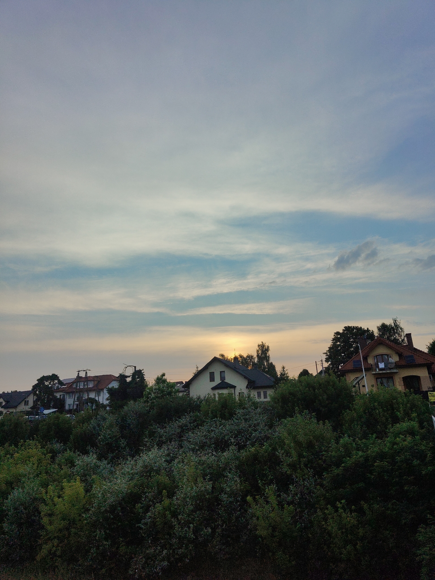 This media file depicts a suburban landscape in Nowy Dwór Mazowiecki, Poland, during what appears to be either sunrise or sunset. The sky is a dominant feature, filled with soft, wispy clouds streaked with pastel hues of blue, white, and a faint yellow or orange glow near the horizon, suggesting the sun is low in the sky.
In the mid-ground, a line of houses peeks out from behind a dense, lush green foliage of trees and bushes that occupies the foreground. The houses exhibit various architectural styles, with pitched roofs in shades of red and dark grey. Some of the houses have solar panels visible on their roofs. A few trees, taller than the surrounding bushes, stand out amongst the residences. Utility poles and wires are also visible, hinting at the presence of infrastructure.
The overall mood is serene and tranquil, with the soft lighting of either dawn or dusk contributing to a peaceful atmosphere. There is no discernible activity or event taking place; it is a static scene of a residential area. The weather appears calm, with no signs of precipitation or strong winds.
A small yellow sign is partially visible on the right side of the image, with some text that appears to read "TAXI" and "MODERN" followed by numbers, suggesting a taxi service is available in the vicinity. The image evokes a sense of quiet suburban life at the edge of the day.