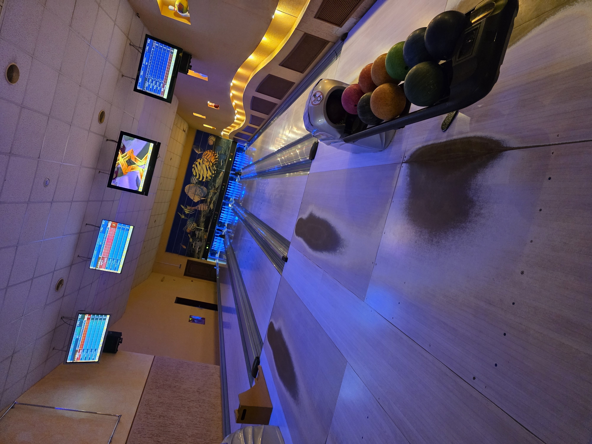 This image captures a vibrant, vertically oriented view inside a modern bowling alley in Chaikovsky, Russia. The scene is dominated by multiple bowling lanes, stretching into the background and illuminated with a strong blue light.
In the immediate foreground, a ball return mechanism is prominent, holding a colorful assortment of bowling balls in shades of green, black, orange, and pink. Scattered on the lane approach area, near the ball return, are visible scuff marks or powder residue, suggesting recent use.
Above the bowling lanes, several flat-screen monitors are suspended from the ceiling. One monitor clearly displays a bowling score sheet, with numbers and a grid typical of a bowling game. Another screen shows an animated golden star against a purple background, possibly an advertisement or game animation. Further down the alley, additional screens are visible with similar score displays.
The ceiling features white, square acoustic tiles with circular recessed lights. Along one side wall, a decorative, wave-like lighting fixture emits a warm yellow glow, complementing the cool blue of the lanes. Below this, a large wall mural depicts stylized fish and aquatic life, adding to the venue's themed decor. The floor in the seating area appears to be a light brown or beige material.
The artificial lighting, including the blue lane lights and warm accents, suggests an evening or nighttime setting. No people are visible, but the setup clearly indicates an active bowling environment. Visible text includes scores and numbers on the screens, typical of a bowling game's interface.