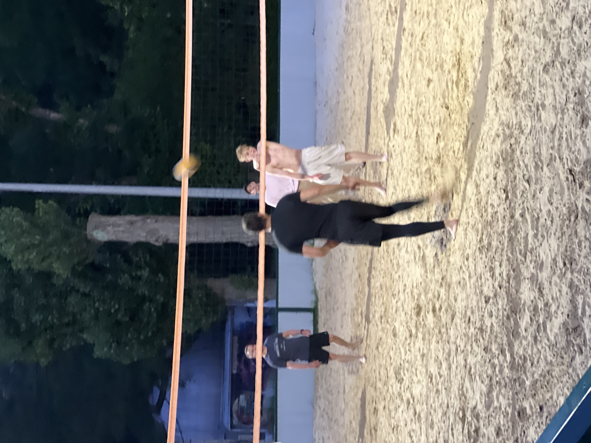 Four men are playing beach volleyball on a sand court at night. The court is illuminated by artificial lights. The men are wearing athletic shorts and t-shirts. Two men are on one side of the net, and two men are on the other side. One man is jumping to hit the ball over the net. The other men are watching the ball or preparing to receive it. The background shows trees and a building. The image was taken in Krasnodar, Russia.