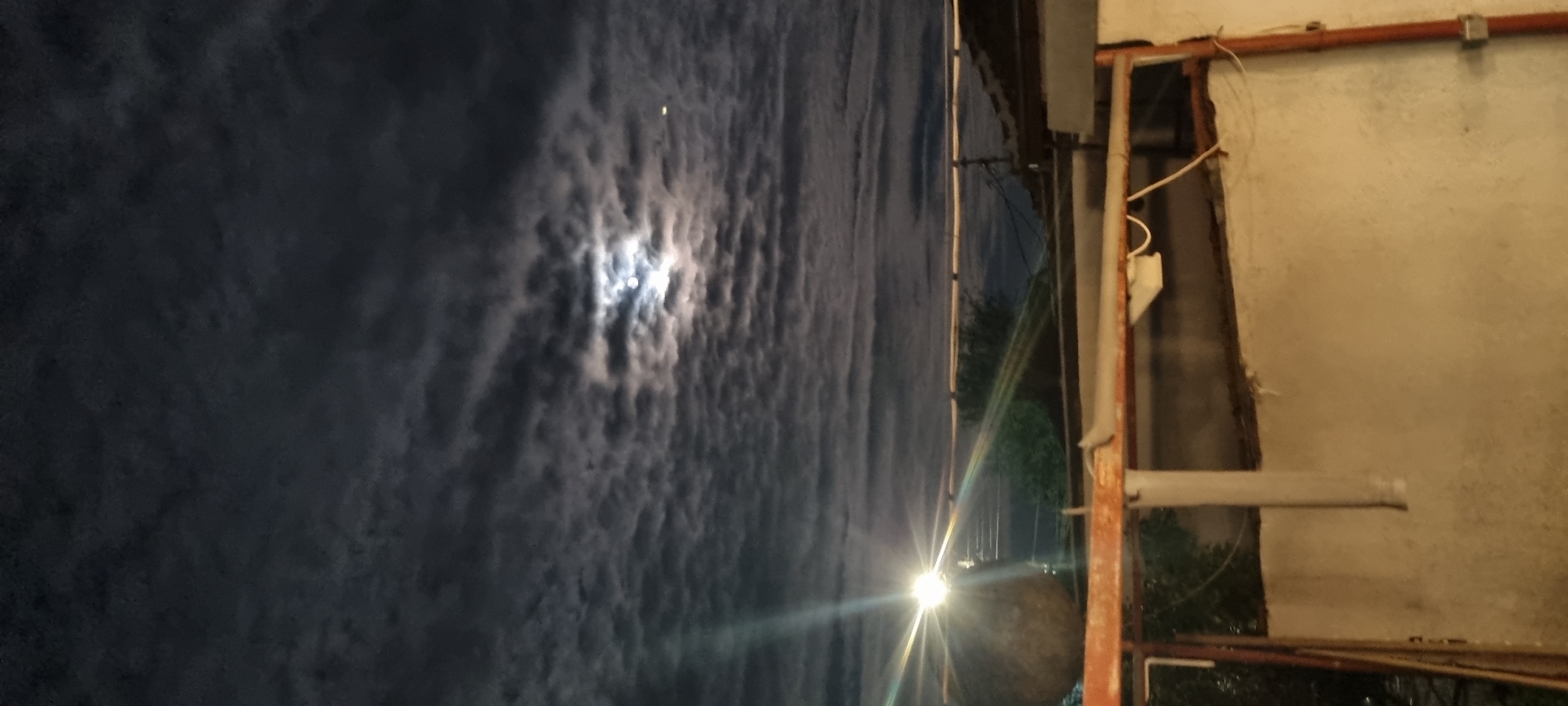 The image depicts a night scene, likely taken from an elevated position in Lom, Bulgaria. The sky is dominated by dark, heavy clouds, partially obscuring a bright moon. A street light or other illumination source is visible in the distance, casting a strong glare. The foreground includes a partially visible building structure with a weathered wall, metal beams, and exposed wiring. A cylindrical object, potentially a tank or container, is present in the middle ground, also illuminated by the distant light. The image is rotated, making the sky appear as a vertical backdrop.
