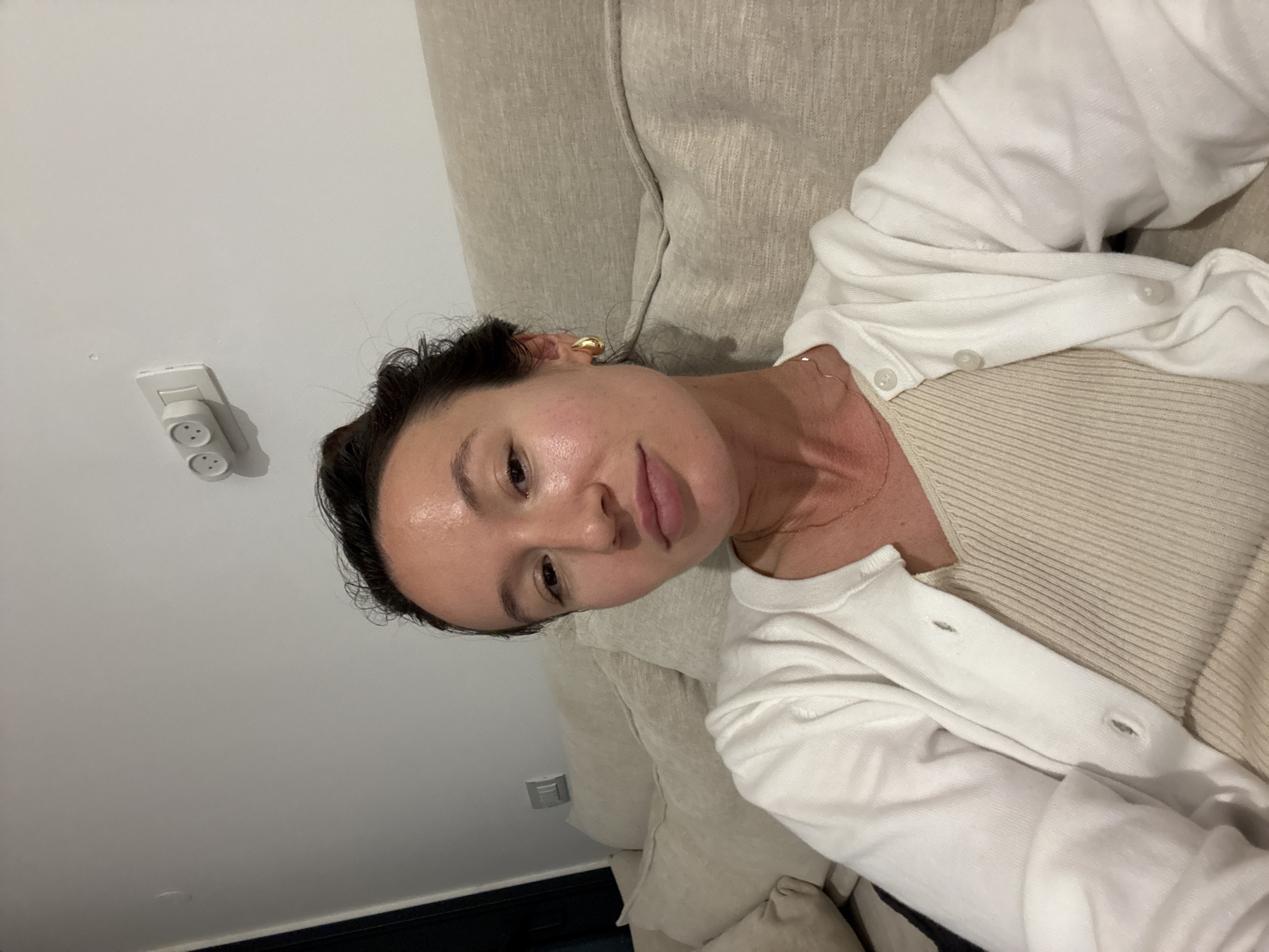 A selfie is taken from the chest up of a woman with dark hair pulled back from her face. She is sitting on a beige couch in front of a white wall. The woman is wearing a white cardigan over a tan ribbed tank top. She has on gold hoop earrings and a thin gold necklace. On the wall behind her, there is a white light switch and two electrical outlets. The scene appears to be indoors, possibly in an apartment or hotel room, though the exact location in Tel-Aviv, Israel cannot be determined from the visual information alone.
