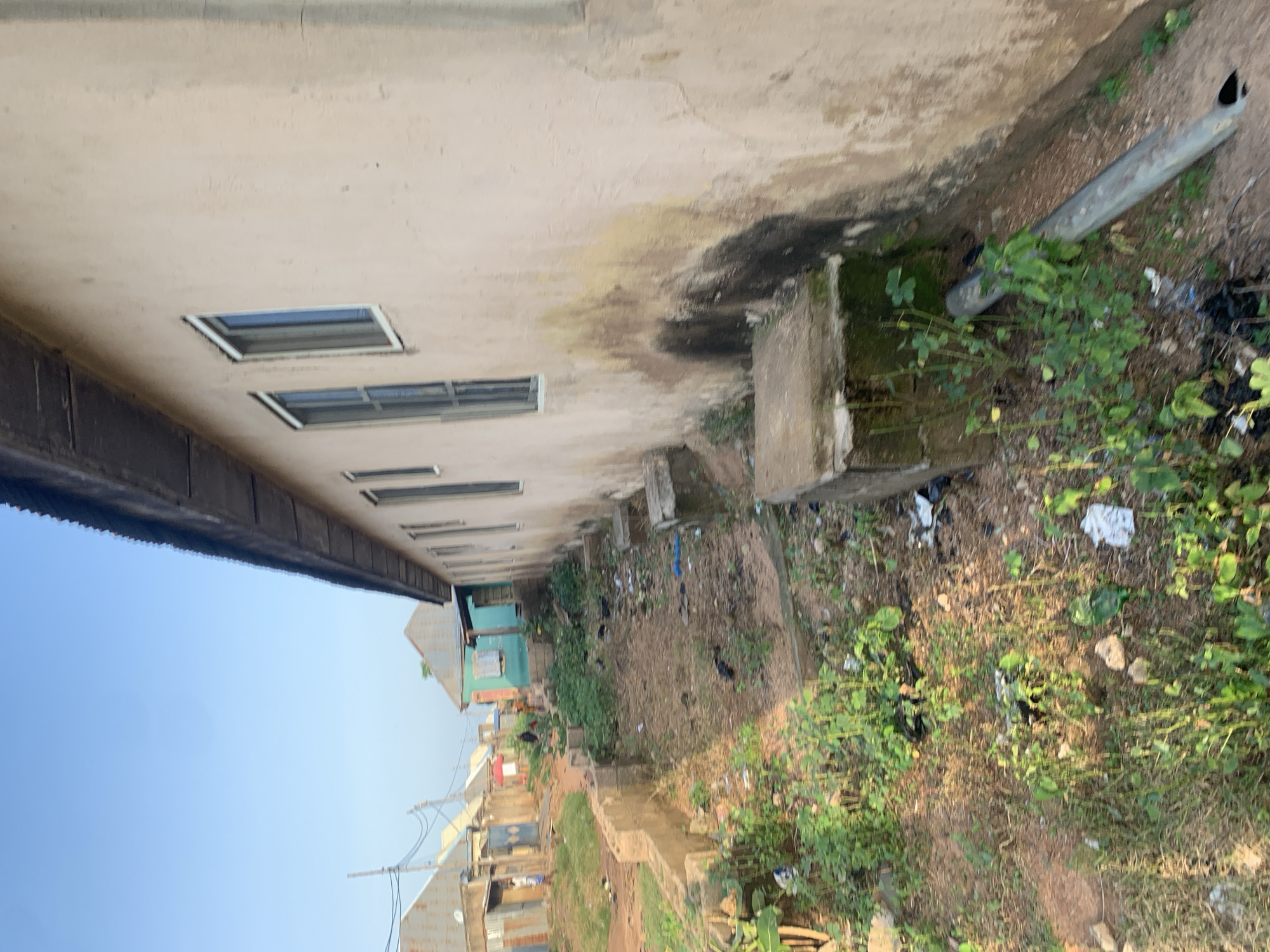 The image depicts an outdoor scene in Jalingo, Nigeria, likely a residential area or a developing neighborhood. In the foreground, a weathered, light-colored building with a dark, corrugated metal roof dominates the right side of the frame. The building has several windows, some with white frames, and the exterior walls show signs of age, with staining and discoloration, particularly dark patches and streaks down the wall. Adjacent to the building, there are concrete structures, possibly foundations or drains, with some vegetation growing around them. The ground is a mix of dirt, scattered debris, and overgrown grass and weeds. Further in the background, other buildings with corrugated metal roofs and varying colors, including blue and green, are visible. A dirt road or pathway runs through the scene, with some people walking in the distance. The sky is clear and blue, suggesting daytime. The overall impression is one of a humble, possibly unfinished, or slightly neglected urban environment.