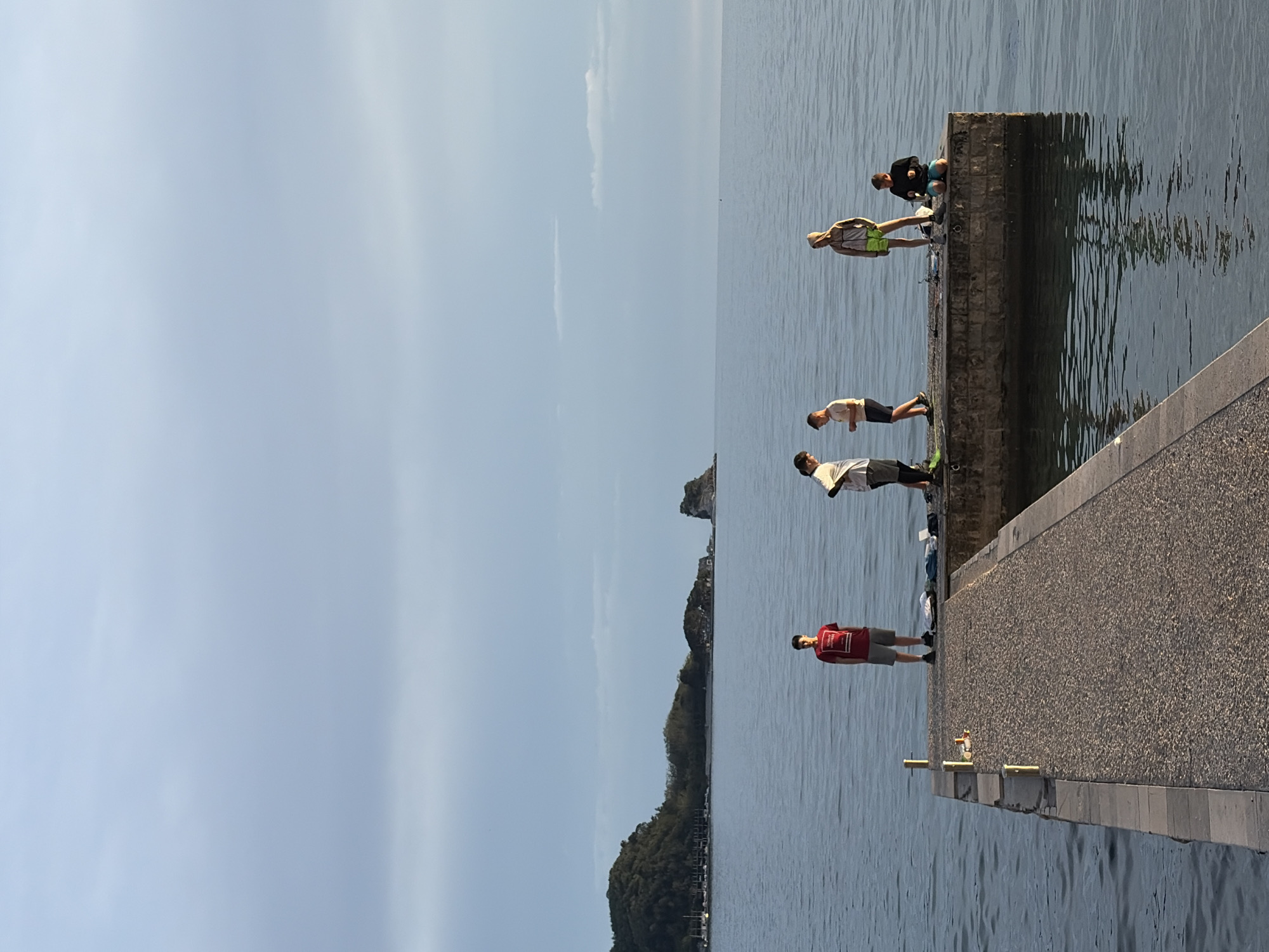 A group of four individuals is positioned along a concrete pier or breakwater extending into the water, under a clear sky. One person, dressed in a red shirt and gray shorts, stands at the start of the pier, facing towards the water. Further along the pier, a person wearing a light-colored top and dark shorts stands near the edge. Adjacent to them is another person with a light-colored shirt and darker shorts. At the pier's end, one individual sits on the structure. The surrounding environment features water with slight reflections. The horizon includes land with buildings. While not explicitly verifiable from the image alone, the scene evokes a coastal location, potentially near Budva, Montenegro.
