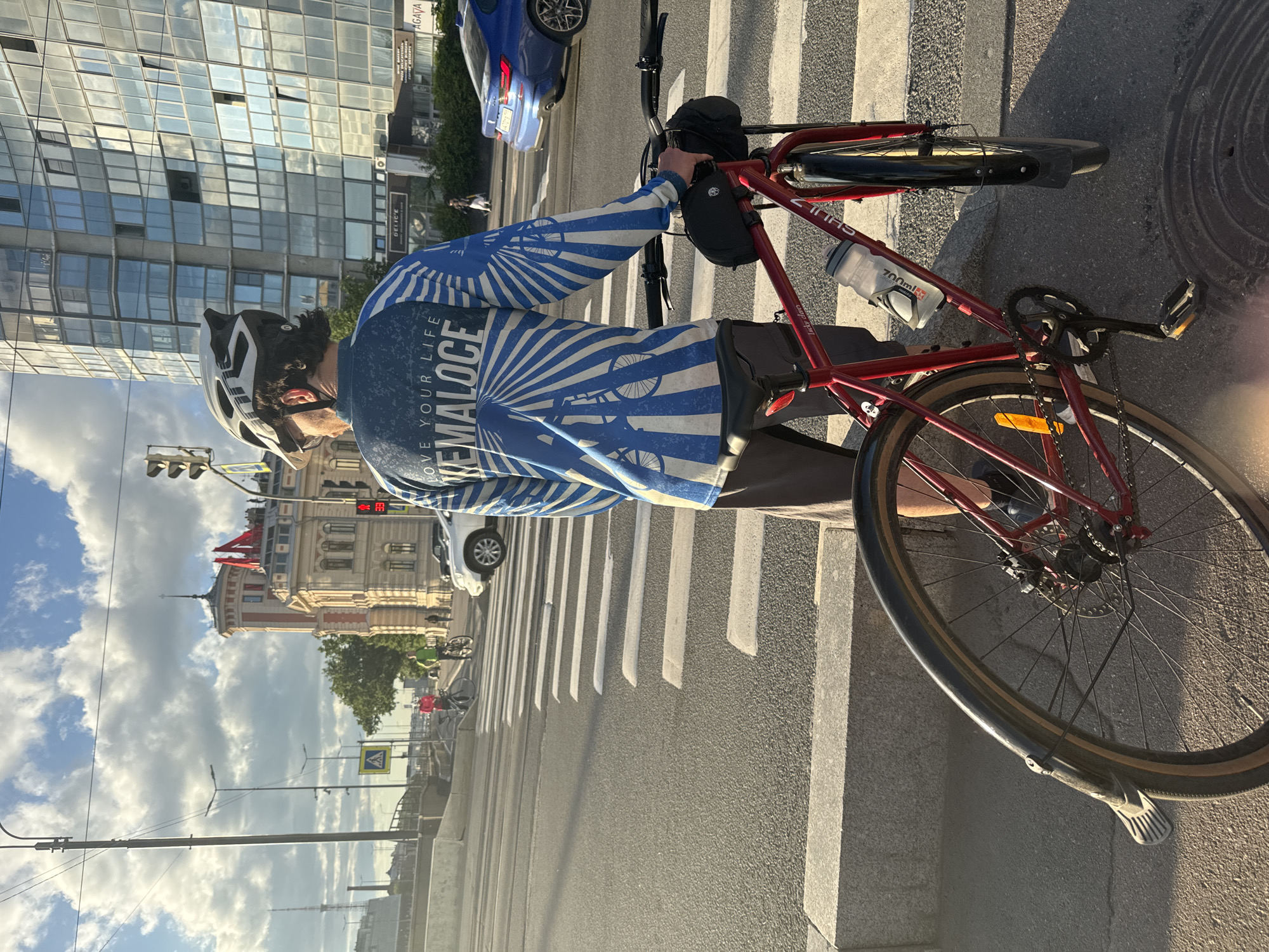A man wearing a cycling helmet and a blue long-sleeved shirt with a white sunburst and bicycle graphic, along with the text "LOVE YOUR LIFE" and "HEMALOCE," stands with his red bicycle at a pedestrian crossing. The bicycle has a black frame bag and a water bottle. The man is on the right side of the image, facing away from the camera and looking towards the street. The scene is set in Saint Petersburg, Russia. In the background, to the left, is a historical building with a tower and red flags. To the right is a modern glass-paneled skyscraper. Traffic lights and street signs are visible. Several cars and other cyclists are present in the background. The sky is partly cloudy. The sun is casting long shadows, indicating it is late afternoon or early evening.