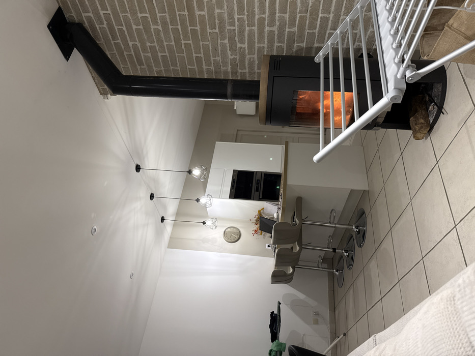 A contemporary interior features a modern black fireplace with a visible fire burning inside, venting through a black pipe attached to a brick wall. A white kitchen island with bar stools sits adjacent to the fireplace. Above the island, three pendant lights hang from the ceiling. A white staircase with metal railings is visible. The flooring appears to be light-colored tile.