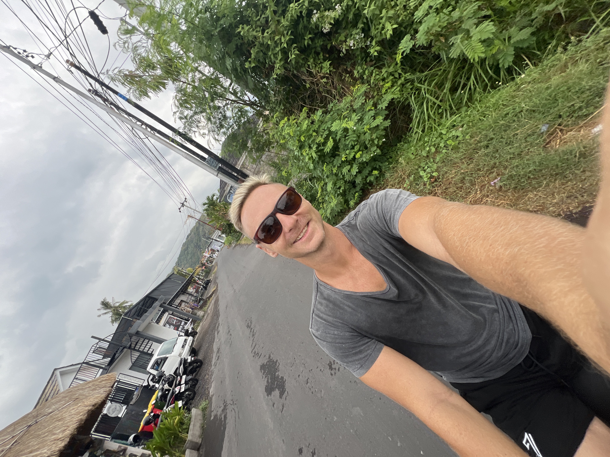 In Abang, Indonesia, a fair-skinned man with blond hair and sunglasses is taking a selfie outdoors. He is wearing a grey t-shirt and black shorts. The man is positioned in the foreground, with the street and buildings in the background. The street is paved and appears to be wet in places. Several motorcycles and a white van are parked along the side of the road. The buildings are of various styles, with some featuring thatched roofs. A dense collection of green foliage lines the right side of the street. Overhead, a complex network of power lines stretches across the cloudy sky.