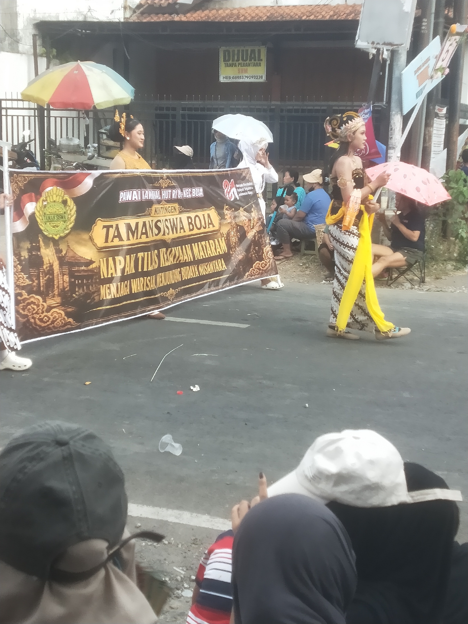 The image depicts a street scene in Boja, Indonesia, during what appears to be a cultural parade or procession. In the foreground, several people are seated on the ground, watching the event. Their backs are to the camera, and they are wearing casual clothing, including a dark baseball cap and a white baseball cap. A large banner stretches across the middle of the image, bearing the text "PAWAI TANHUL HUT RI & KEC BOJA" and "TAMANSISWA BOJA NAPAK TILAS KERAJAAN MATARAM MENJAGA WARISAN BUDAYA NUSANTARA." The banner features an illustration of traditional Indonesian architecture.
To the right of the banner, two women are participating in the procession. One woman, dressed in traditional attire with an elaborate headdress, holds a pink umbrella and stands to the right. Another woman in a white headscarf and traditional clothing is also visible. Behind them, other participants and spectators can be seen, along with a few umbrellas and a building in the background that has a "DIJUAL" (For Sale) sign. The street is paved, and there are some scattered debris on the ground. The overall atmosphere suggests a festive community event.