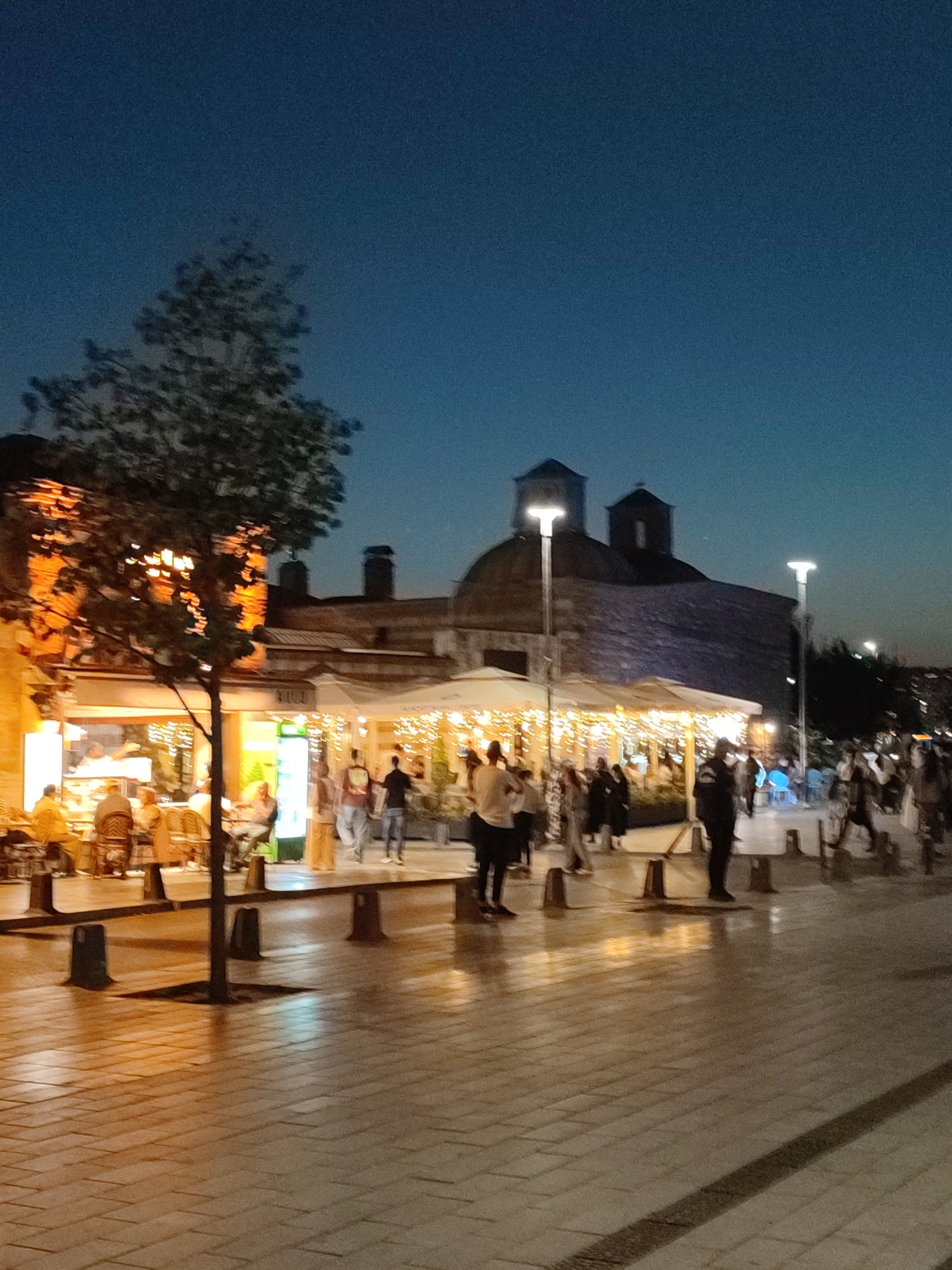 The image captures a street scene at dusk in Üsküdar, Turkey, likely in the vicinity of the Maiden's Tower. In the foreground, a tree stands on a paved walkway, its silhouette stark against the illuminated buildings and streetlights. The walkway is made of rectangular pavers and reflects the warm glow of the lights. Several people are visible, some walking and others seated at outdoor tables of a cafe or restaurant. The establishment is illuminated by strings of lights and features a bright green refrigerator. In the background, a historic building with a dome and two cupolas is partially visible, its facade weathered and aged. Streetlights line the walkway, casting pools of light and illuminating the scene. The sky is a deep twilight blue, indicating the transition from day to night. The overall atmosphere is one of urban leisure, with people enjoying the evening outdoors.