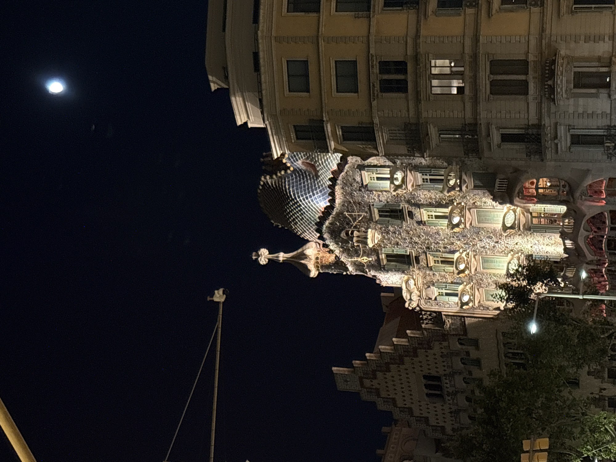 This image depicts Casa Batlló, a building designed by Antoni Gaudí, located on Passeig de Gràcia in Barcelona, Spain. It is nighttime, and the building is illuminated, highlighting its distinctive organic and colorful facade. The moon is visible in the dark sky, along with some stars and a distant airplane or satellite. The building's architecture features a wavy roof covered in mosaic tiles resembling scales, and balconies that appear to be skeletal. The facade is textured and appears to be made of stone, with irregularly shaped windows and decorative elements. Adjacent buildings, also illuminated, are visible on either side, one with a more traditional, angular design and another with a lighter, modern appearance. Streetlights and trees are also present in the foreground, suggesting an urban street scene. The overall impression is a surreal and fantastical architectural marvel against a night sky.