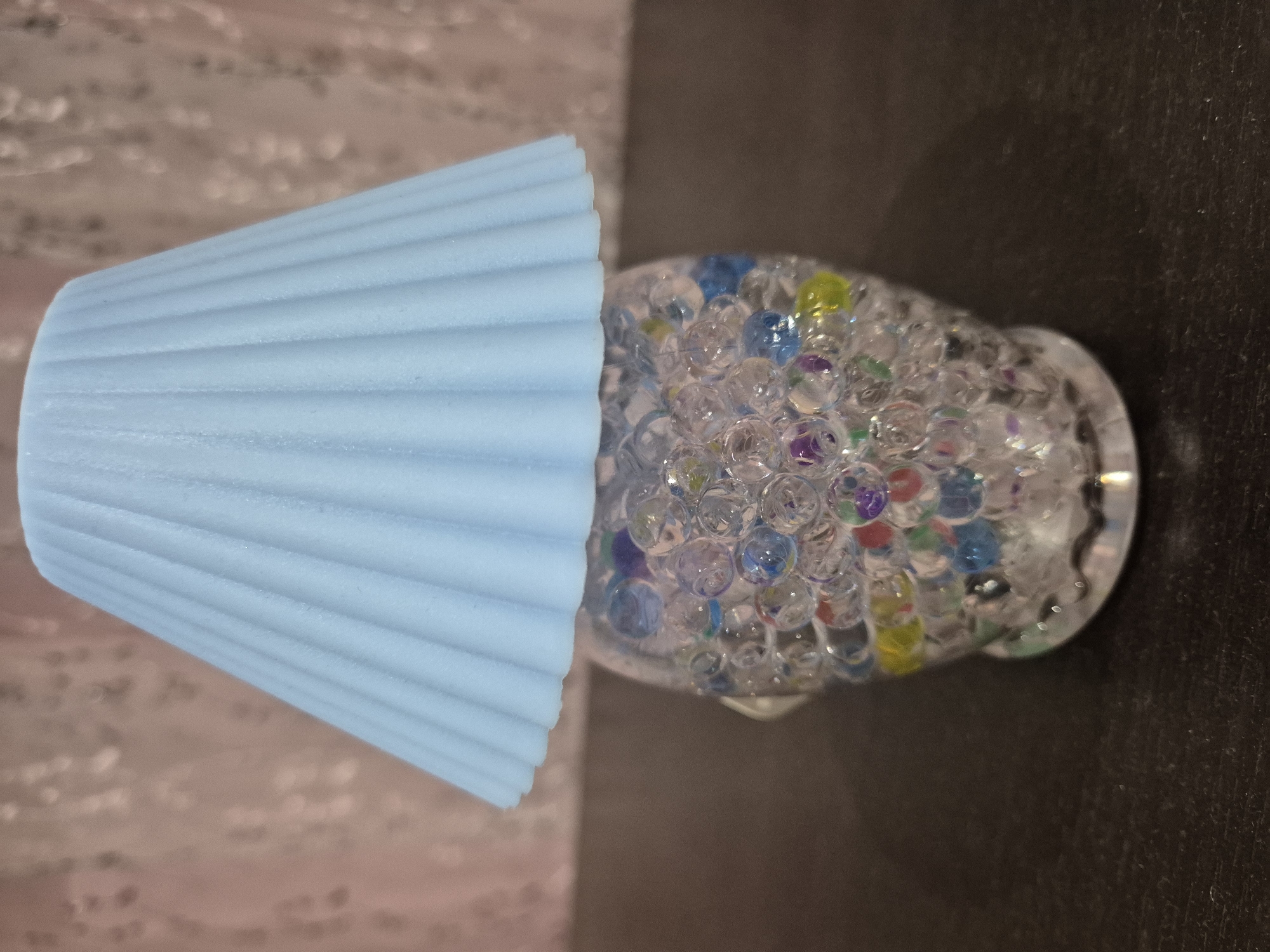A light blue, ribbed, cone-shaped object is adjacent to a clear glass container filled with hydrogel beads of various colors, including blue, purple, red, and yellow. The objects are on a surface, the left side of which has a light-colored, textured pattern, and the right side is a dark, solid color. The location and city are unknown.