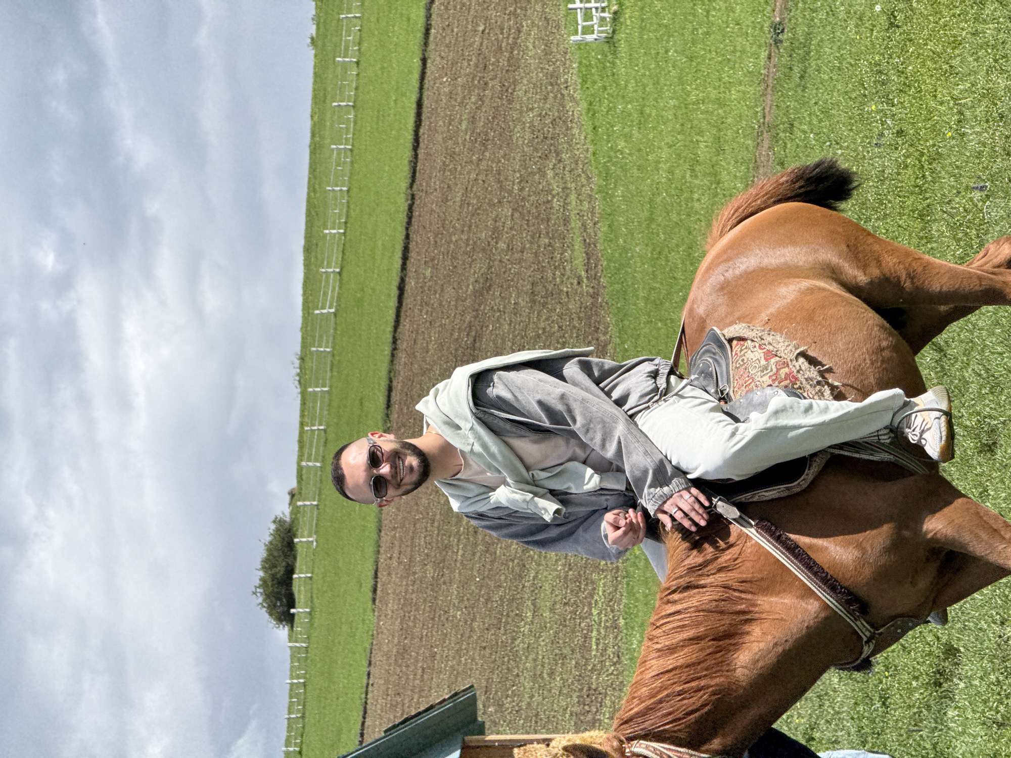 A man is sitting on a chestnut horse outdoors. The man wears sunglasses, light-colored jeans, a light-colored shirt, and a gray jacket tied around his shoulders. The horse is equipped with a saddle and bridle. The setting appears to be a grassy field with a fenced agricultural area in the background, possibly in the city of Dsegh, Armenia. The sky is overcast.