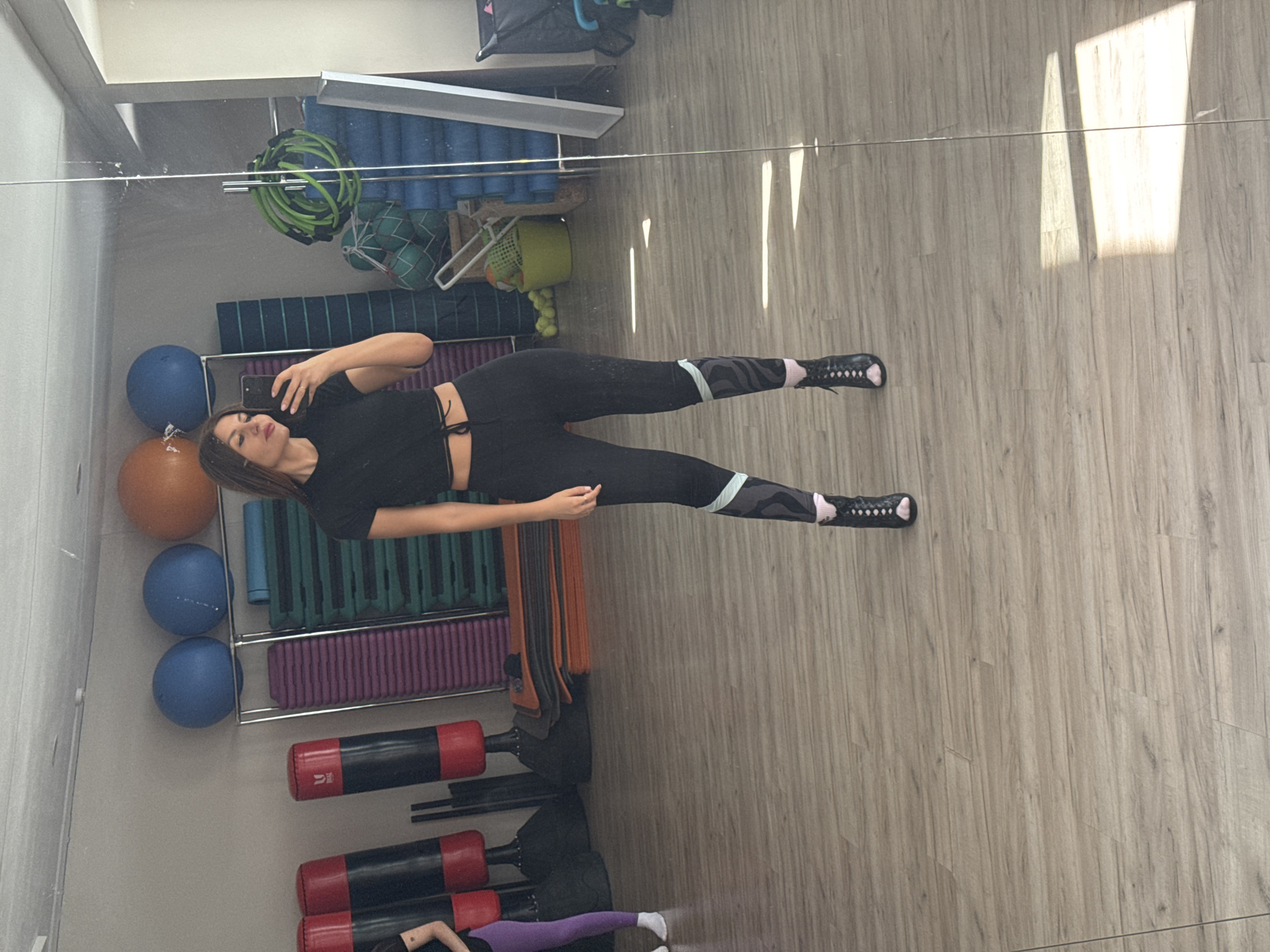 A woman in black athletic wear is taking a selfie in a mirror. She is standing in a gym in Chaikovsky, Russia. The gym has exercise balls, mats, punching bags, and other gym equipment. The woman has dark hair. She is wearing black leggings and a black crop top. The leggings have a stripe near the ankle. She is holding a cell phone in her left hand.