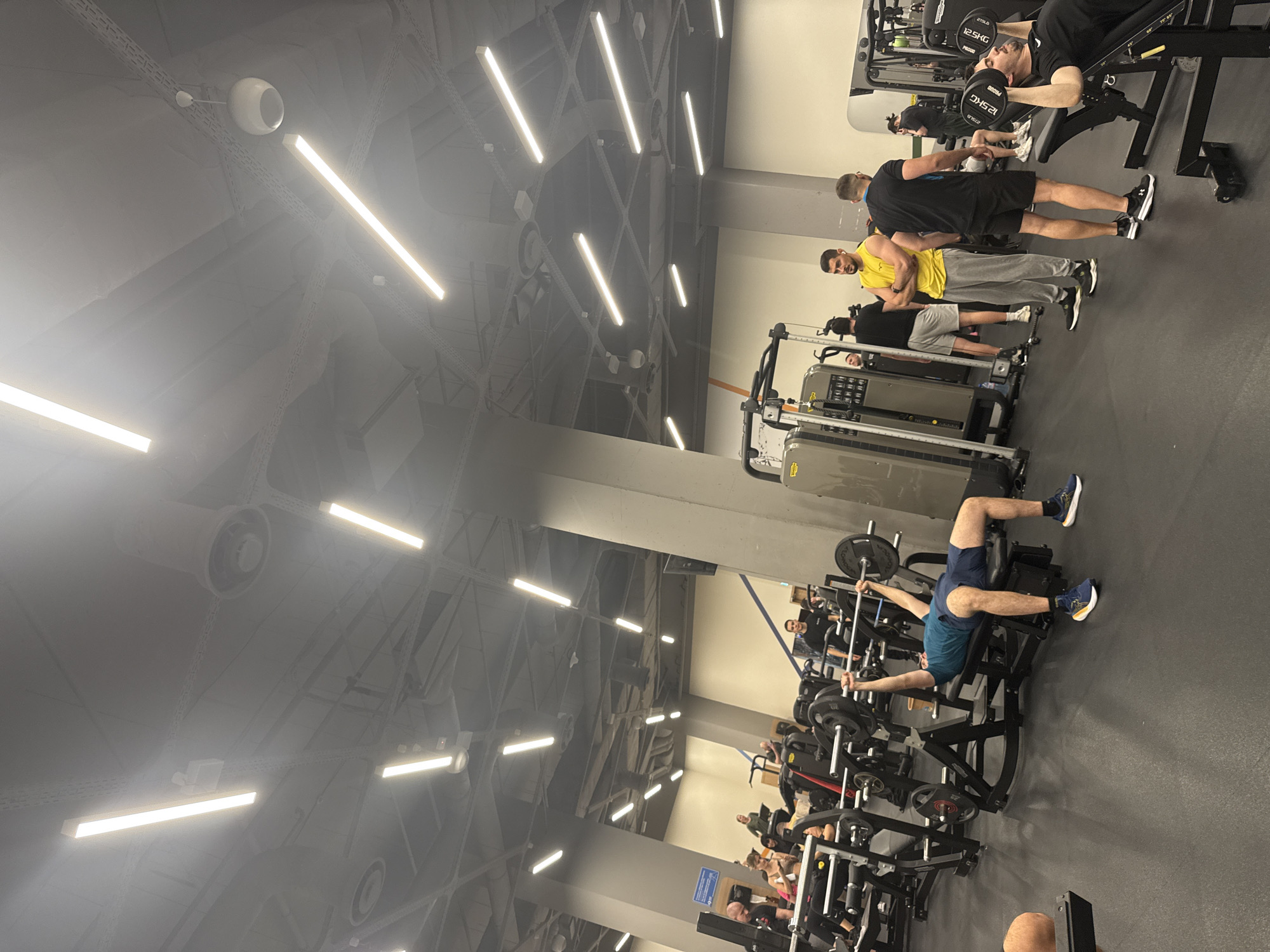 The image captures a gym scene, possibly in Sofia, Bulgaria. Several individuals are actively engaged in various exercises using weight machines and free weights. The gym is well-lit with linear ceiling lights and ventilation systems. The floor is a consistent dark grey. There is a mix of men, some wearing workout gear such as shorts and t-shirts. People are focused on their individual workouts. Some are lifting weights while lying on benches, others are using machine-assisted weightlifting equipment. The environment is modern and appears well-maintained.