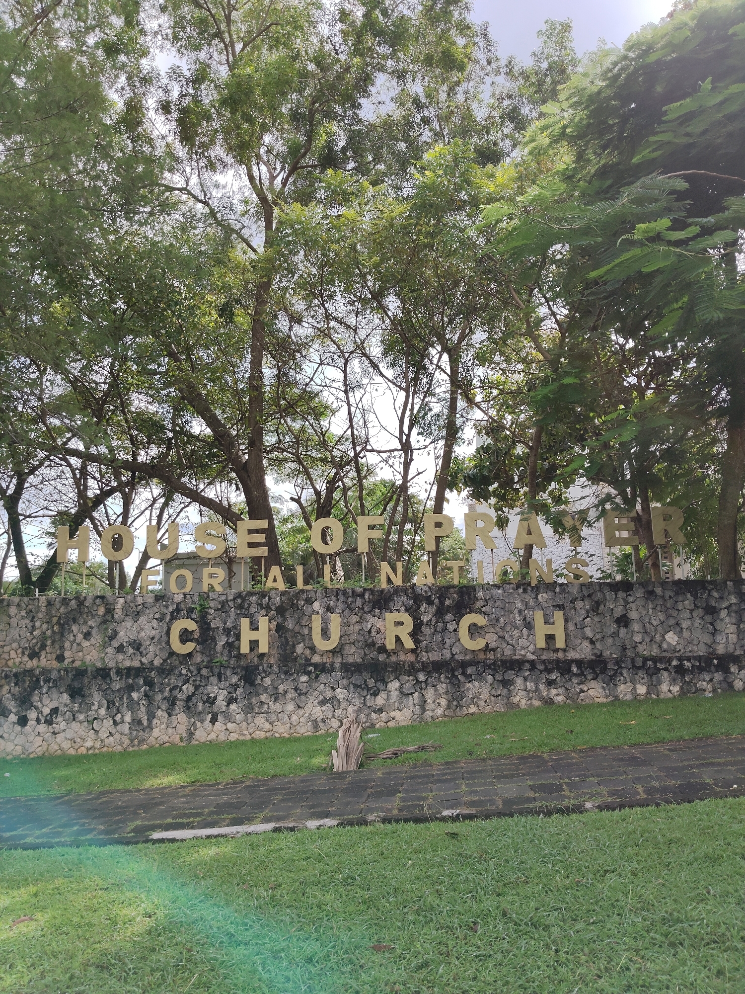 This image captures the entrance to the "House of Prayer for All Nations Church" located in Ungasan, Indonesia. The church's name is displayed in large, gold lettering affixed to a stone wall. A patch of green grass extends from the base of the wall towards the foreground. The scene is framed by a backdrop of trees with dense foliage. The sky is overcast.