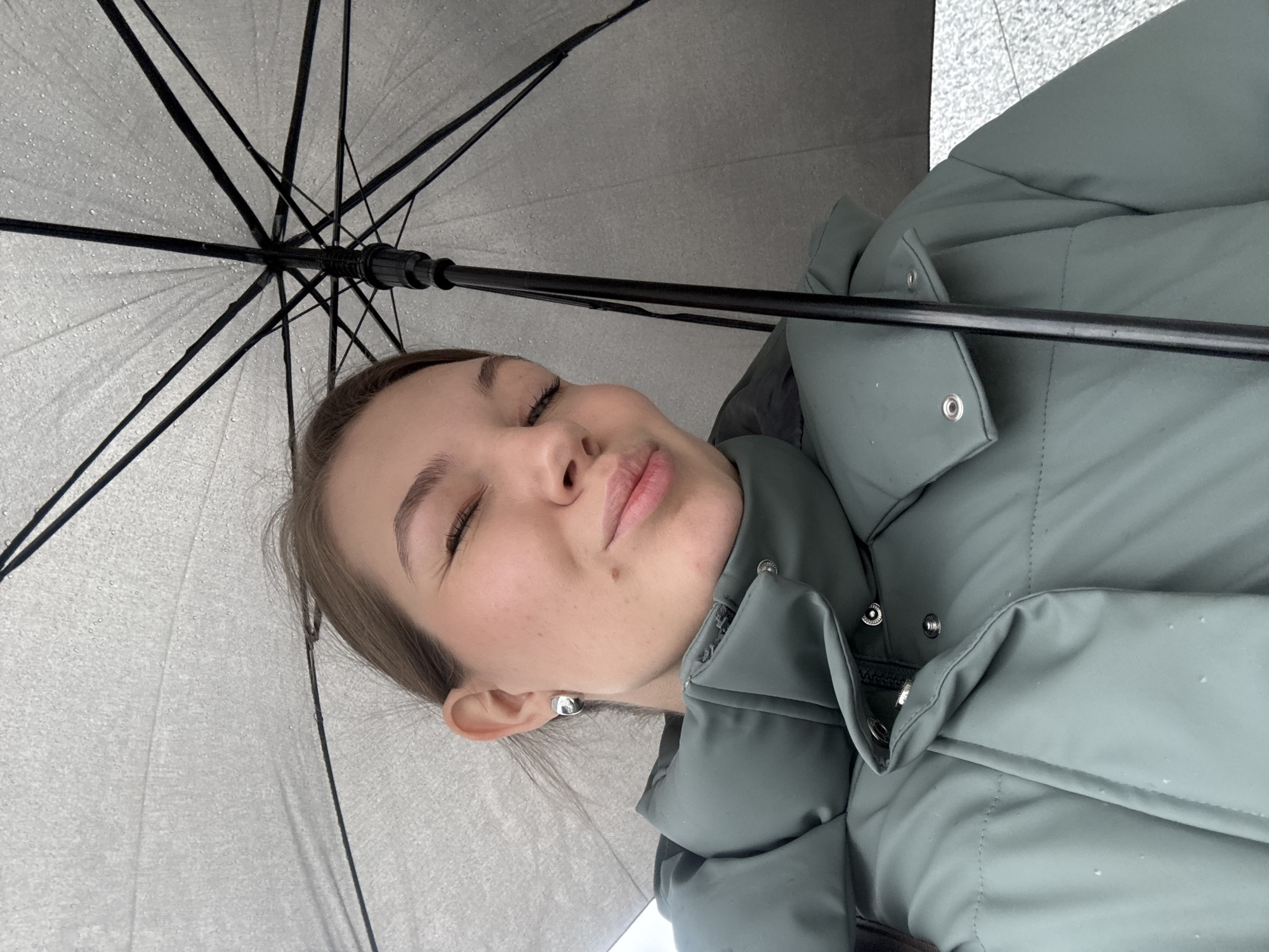 A woman with light skin and a slight smile poses under an umbrella. She wears a gray-green puffer jacket and silver stud earrings. The umbrella above her is a light gray color and water droplets are visible on its surface. The background is blurred and indistinct, but appears to be an outdoor setting, possibly a street, with hints of gray concrete. The location is Belgrade, Serbia.