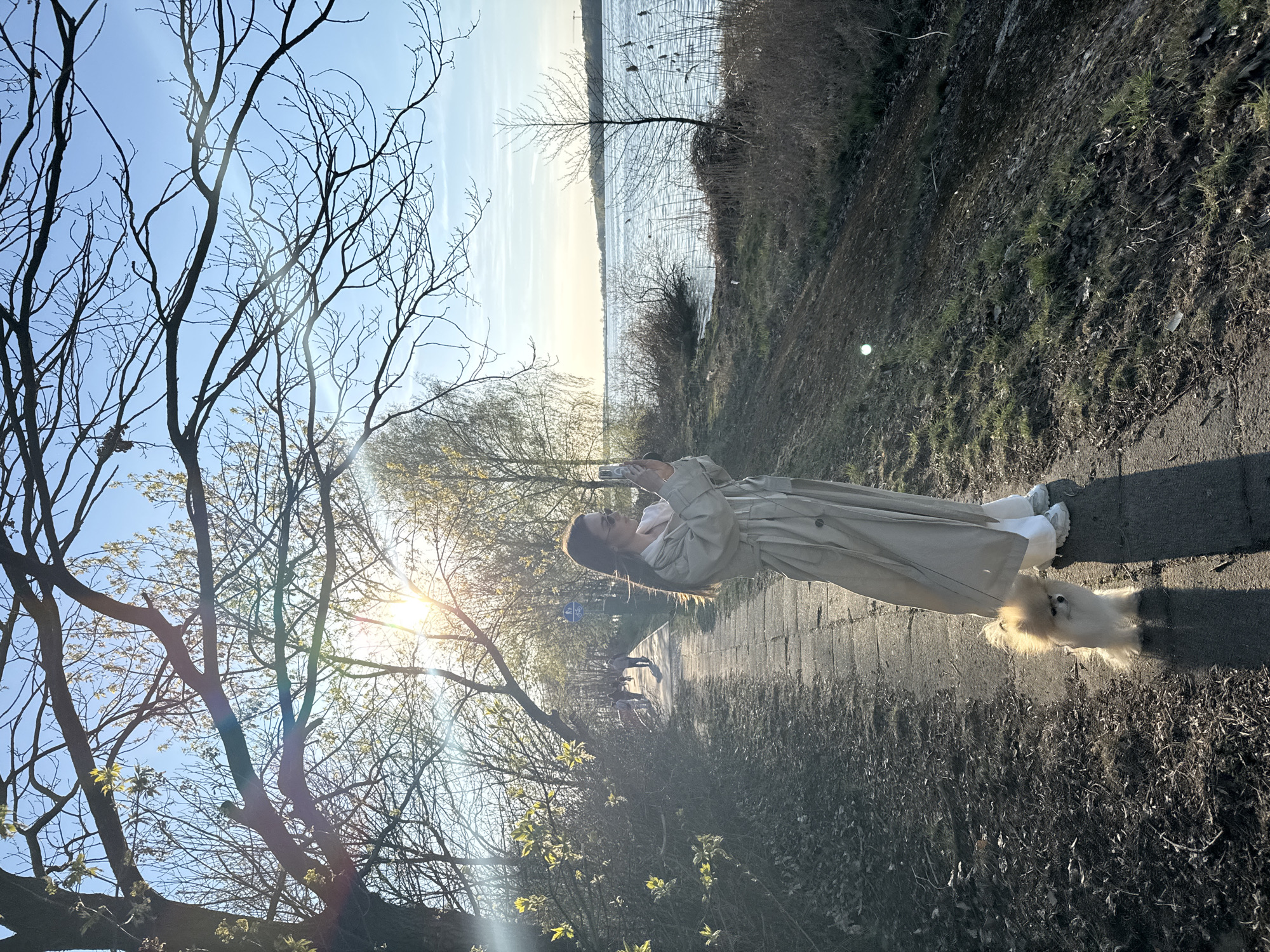 A woman in a beige coat and white pants stands on a paved path next to a fluffy white dog. She holds a phone up, possibly taking a picture. To the left are bare trees with the sun shining through them. In the background, a body of water is visible. The location is Białobrzegi, Poland.