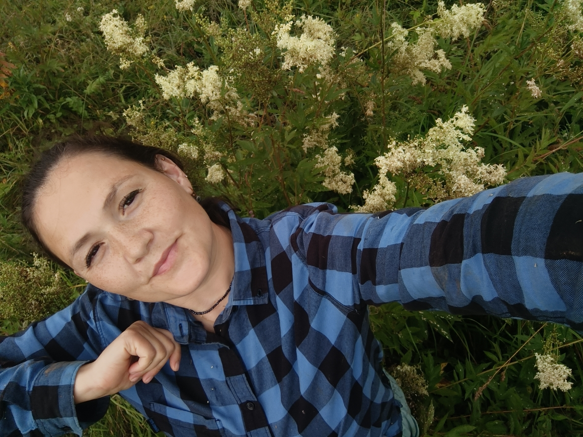 A medium shot captures a young woman with dark hair and light brown eyes lying in a field of white wildflowers and green grass, taking a selfie. She is wearing a blue and black checkered long-sleeved shirt and a dark beaded necklace. Her left arm is bent, and her hand rests on her chest. Her face is turned towards the camera, and she is smiling slightly. The background consists of lush green foliage and clusters of small white flowers. The lighting suggests an outdoor setting, possibly during the daytime.