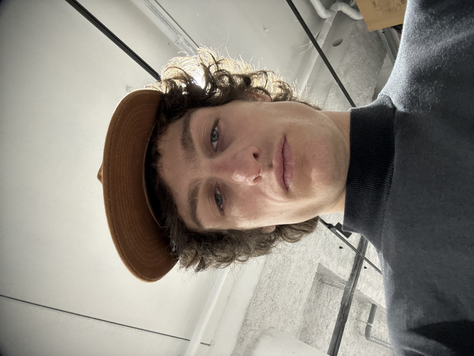 A male individual is depicted in a low-angle, close-up photograph taken indoors in Joinville-le-Pont, France. The subject, appearing to be an adult male, has curly dark brown hair and light-colored eyes, with a small metallic ring piercing on the right nostril. He wears a black mock neck shirt and a flat-brimmed, brown-toned hat resting atop his head. His expression is neutral, and he is looking directly towards the camera. The background reveals an interior space with a light-colored, textured ceiling, featuring dark linear structural elements or tracks, and white pipes. Illumination appears to be overhead, creating a backlight effect on the subject's right side.