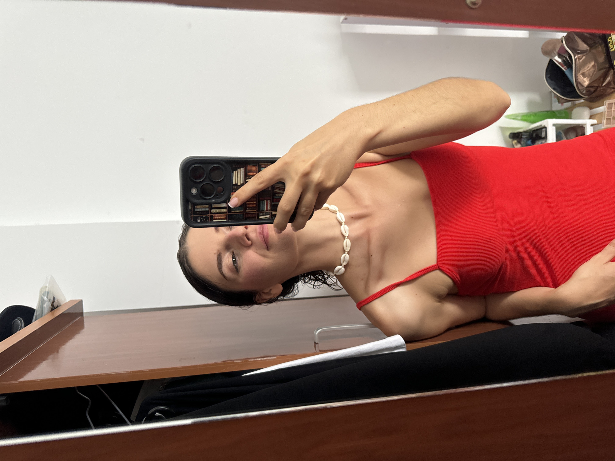 An indoor mirror selfie captures a young woman looking directly at the camera with a subtle smile. She has dark hair, which appears slightly wet or pulled back, and is wearing a vibrant red, ribbed top or dress with thin straps. Around her neck, she sports a white choker necklace made of seashells.
She holds a black smartphone with multiple camera lenses in her right hand, positioned to reflect herself in the mirror. The phone is encased in a distinctive cover designed to resemble a full bookshelf, featuring numerous miniature book spines.
The reflection in the mirror reveals a brown wooden surface, possibly a desk or vanity, in the lower foreground. The background behind her consists of a plain white wall, and to her right, a cluttered shelf holds various personal items. These include a dark bag with a metallic finish (possibly a makeup or toiletry bag), a clear plastic container, and other indistinct objects, suggesting a personal living space. The provided context indicates the location is Mũi Né, Vietnam. No discernible text is clearly readable within the image, apart from the decorative book spine designs on the phone case.