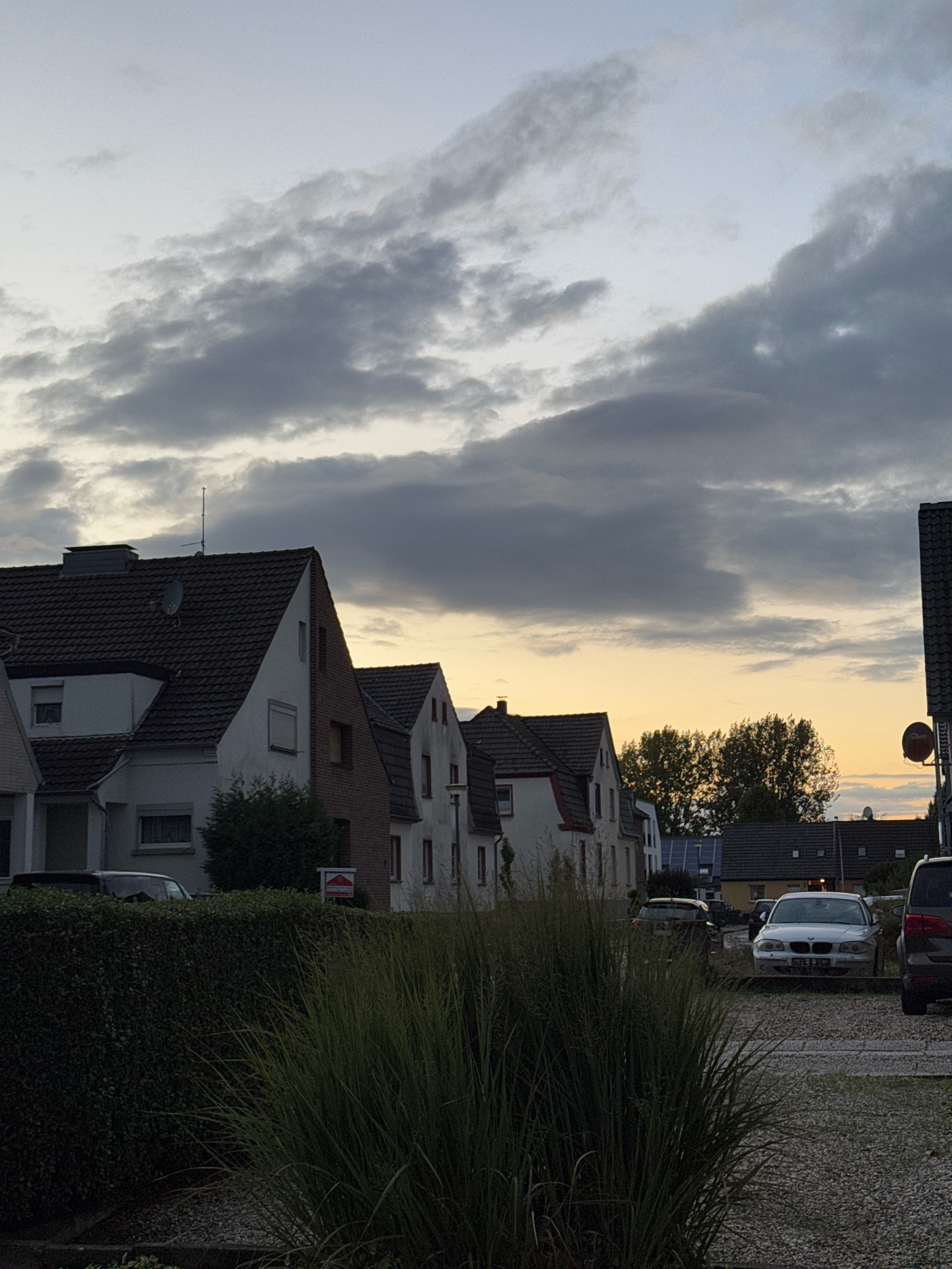 The image depicts a street scene in the city of Cleves, Germany. The sky is overcast with dramatic clouds. In the foreground, there is a dense green hedge and tall grass. Behind the hedge, a row of attached houses with tiled roofs is visible. Some of the houses have white facades, while others are made of red brick. Several cars are parked on a gravel driveway in front of the houses. Further in the background, there are trees and more houses, suggesting a residential neighborhood. The lighting indicates it is either early morning or late evening.