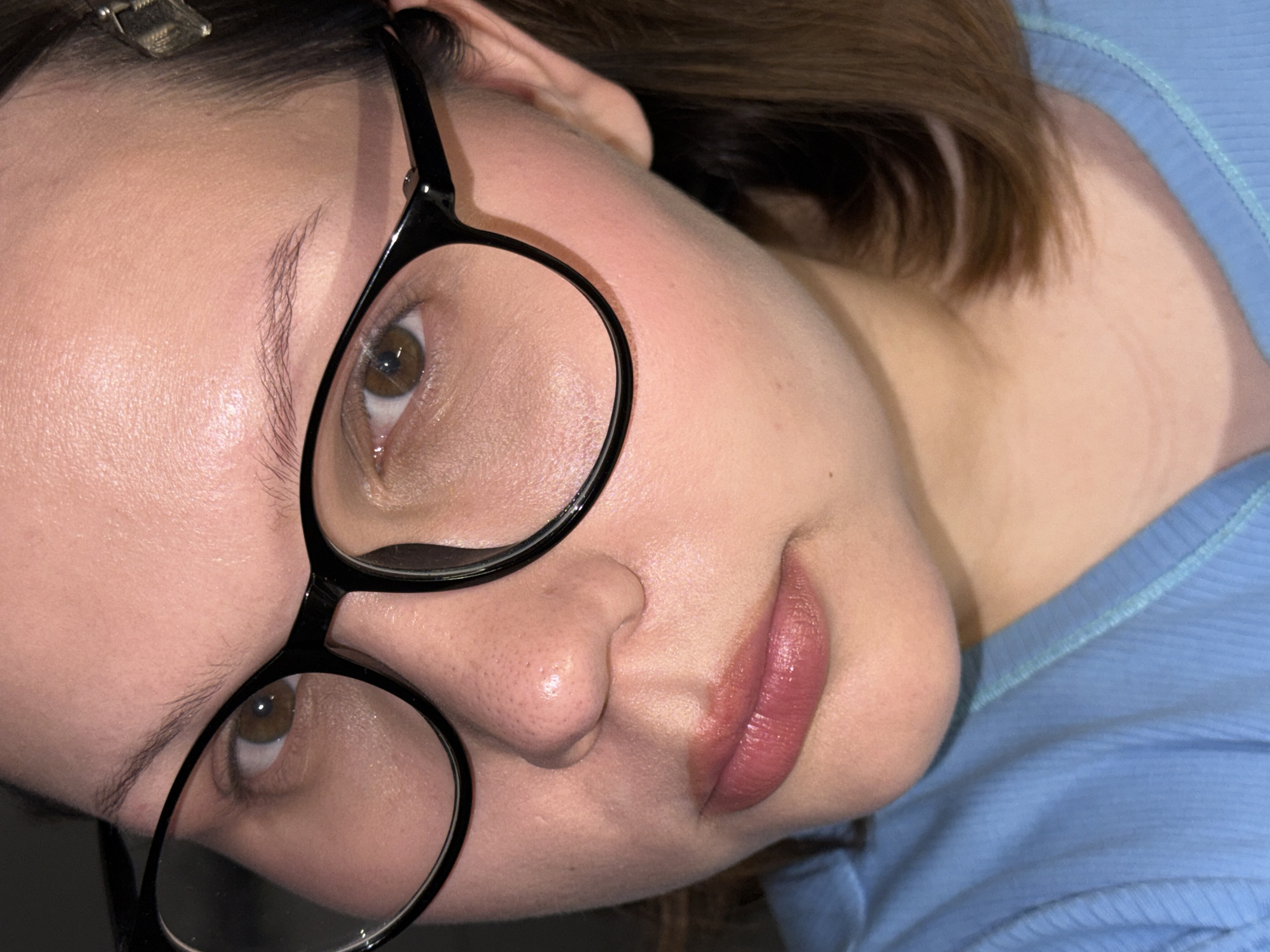 A close-up photograph depicts a person's face, angled towards the camera. The person is wearing black, round-framed glasses. The face appears to be naturally lit, showcasing skin texture details. The individual's lips are tinted with a nude-pink color. A blue shirt is visible on the right side of the frame. There is a hair clip in the upper-left area of the picture. There is no information that indicates the picture's location is in Chaikovsky, Russia.