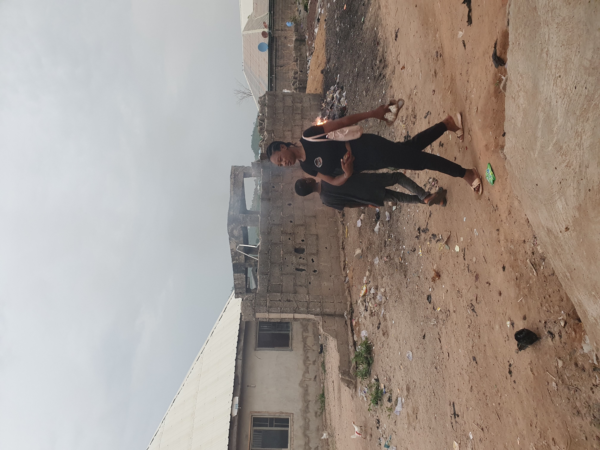 This image, captured in Lokoja, Nigeria, depicts an outdoor scene featuring two individuals engaged in an activity involving a sparking object. The setting appears to be an informal settlement or a developing area characterized by unpaved ground and rudimentary structures under an overcast sky.
In the foreground, a young woman, dressed in a black t-shirt with a white peace symbol, black pants, and light-colored flip-flops, stands holding a light-colored shoulder bag. She is holding an object in her right hand that is actively emitting bright, glowing sparks, resembling a sparkler or small firework. Her attention is directed towards this sparking object. Facing her is a younger individual, likely a child, also dressed in dark clothing (t-shirt and pants) and dark flip-flops. This person's hands are raised near the sparking object, suggesting interaction or observation.
The ground beneath them is a reddish-brown dirt or sandy surface, heavily strewn with trash including plastic bags, wrappers, and general debris. In the background, several buildings constructed from concrete blocks are visible. Some structures appear to be in various stages of construction or disrepair, with exposed blockwork and broken sections. One building has a light-colored corrugated metal roof, while another features dark-paned windows. A small white bird, possibly a chicken, can be seen on the ground near one of the buildings. The sky is uniformly light grey and overcast, indicating daytime. The scene conveys a casual moment of play or celebration amidst the everyday environment.