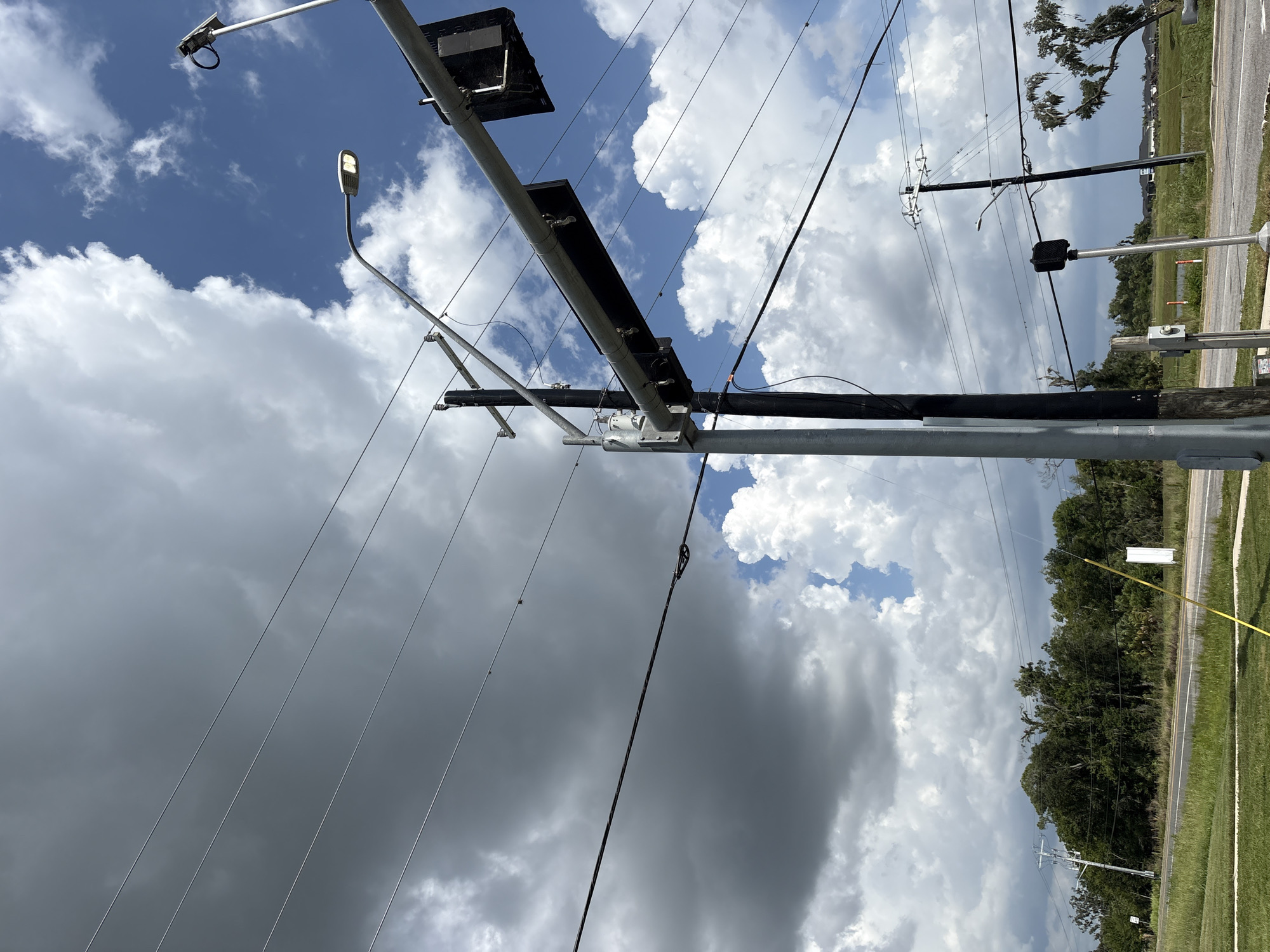 The image depicts an outdoor scene in Parrish, United States, dominated by a cloudy sky with patches of blue. In the foreground and mid-ground, several utility poles and power lines are visible, stretching across the frame. One pole, a prominent vertical element, supports traffic signals and a street light. The traffic signals are black and rectangular, and the street light is illuminated. Another utility pole further back also carries power lines and a street light. The ground in the foreground is covered with green grass, and a road is visible in the mid-ground. Beyond the road, there is a line of dense trees. In the background, residential structures and a body of water can be seen. The overall impression is that of a street scene on a partly cloudy day.