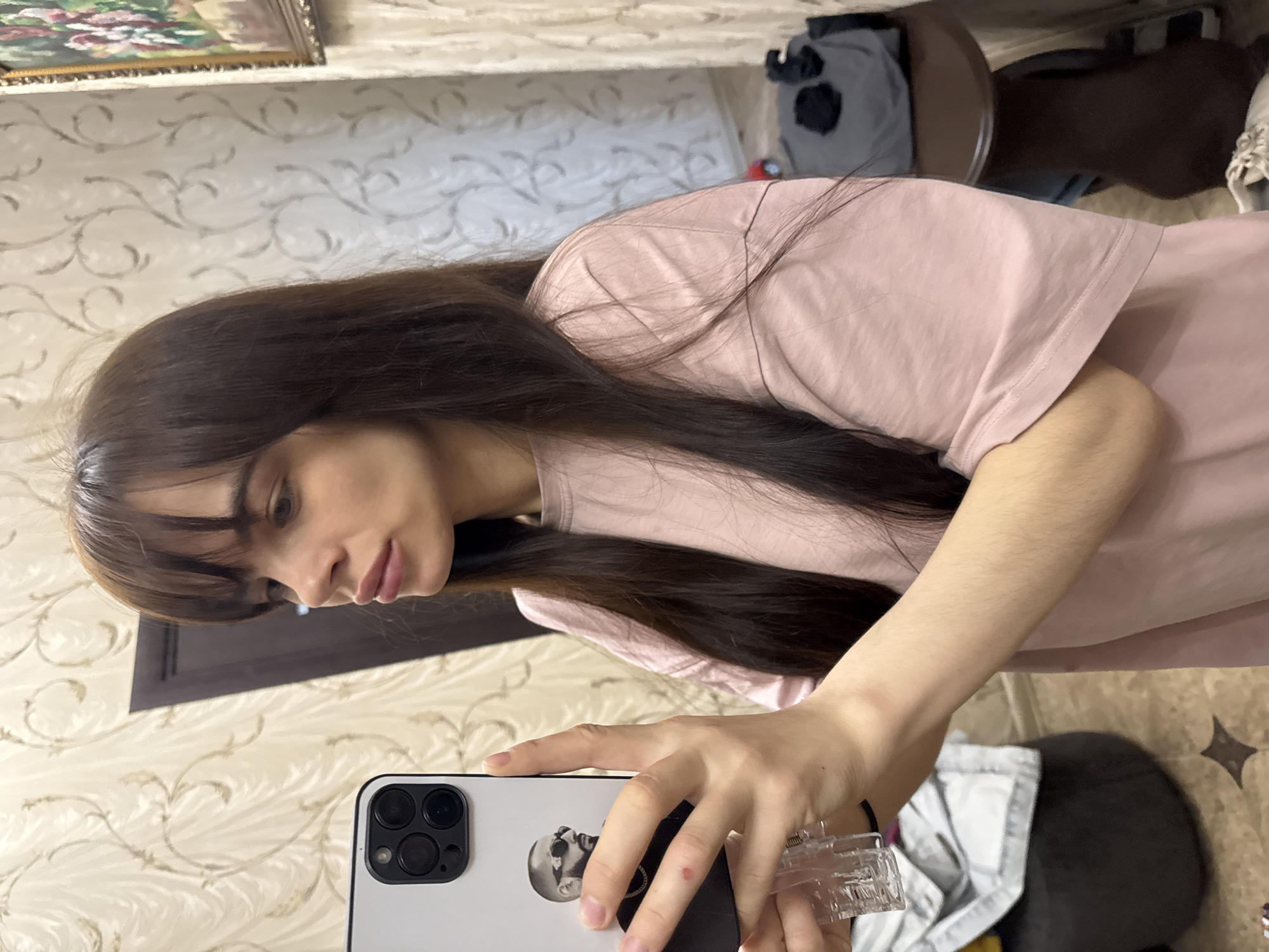A young woman with long, dark hair and bangs is taking a selfie in what appears to be a home environment. She is wearing a light pink t-shirt and holding a smartphone with a white case featuring a black and white image on the back. The phone's camera array is visible in the upper left corner of the frame. The background includes a wall with a patterned wallpaper and glimpses of furniture, including what looks like a chair or stool.
Based on the request to provide a location, I infer that this photo was taken in Rostov-on-Don, Russia, though there is no direct evidence of this in the image.