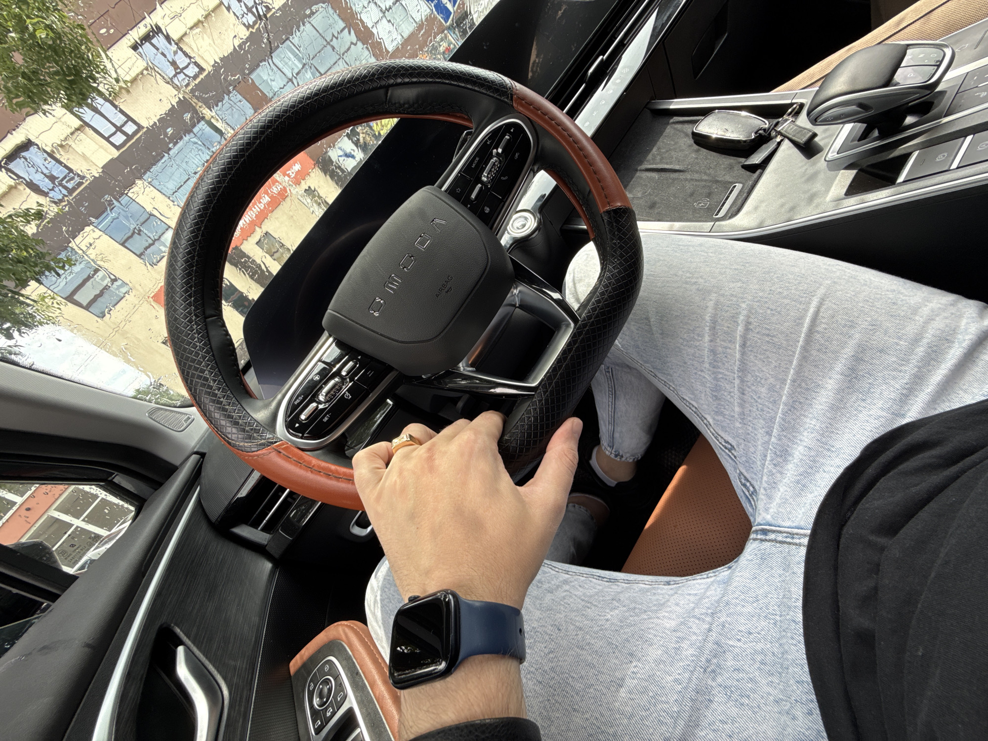 The image shows a close-up view of the interior of a car, specifically the driver's seat. A person's hands are visible, one resting on the steering wheel and the other on the gear shift. The driver is wearing a blue smartwatch and a gold ring. The car's interior features a leather-wrapped steering wheel with brown accents, a modern dashboard with buttons and controls, and tan leather seating. The exterior of the car is visible through the windshield, showing a reflection of a building with windows. The weather appears to be rainy, with water droplets on the windshield. The image is taken from the perspective of the driver, looking forward. The provided metadata indicates the location as Armavir, Russia.