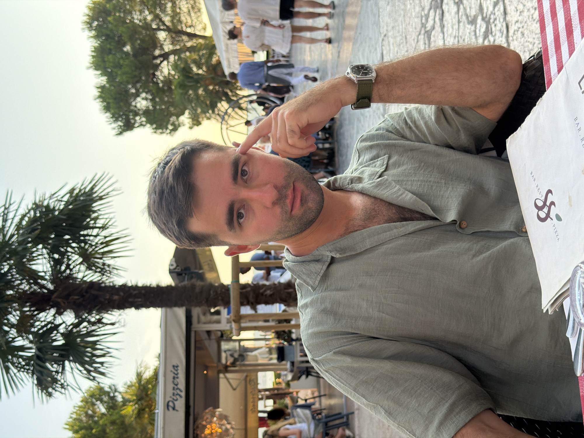 A man with short brown hair and a light beard is sitting at a table outdoors, with his left hand resting on his forehead. He is wearing a light green, open-collar, short-sleeved shirt. A watch with a green band is visible on his left wrist. In front of him is a menu with red and white checkered tablecloth. Behind him, a palm tree stands tall, and further back, there's a restaurant with a "Pizzeria" sign. Other people are visible in the background, walking near the waterfront. The scene appears to be set in Sirmione, Italy, likely during the late afternoon or early evening, given the soft, warm lighting.