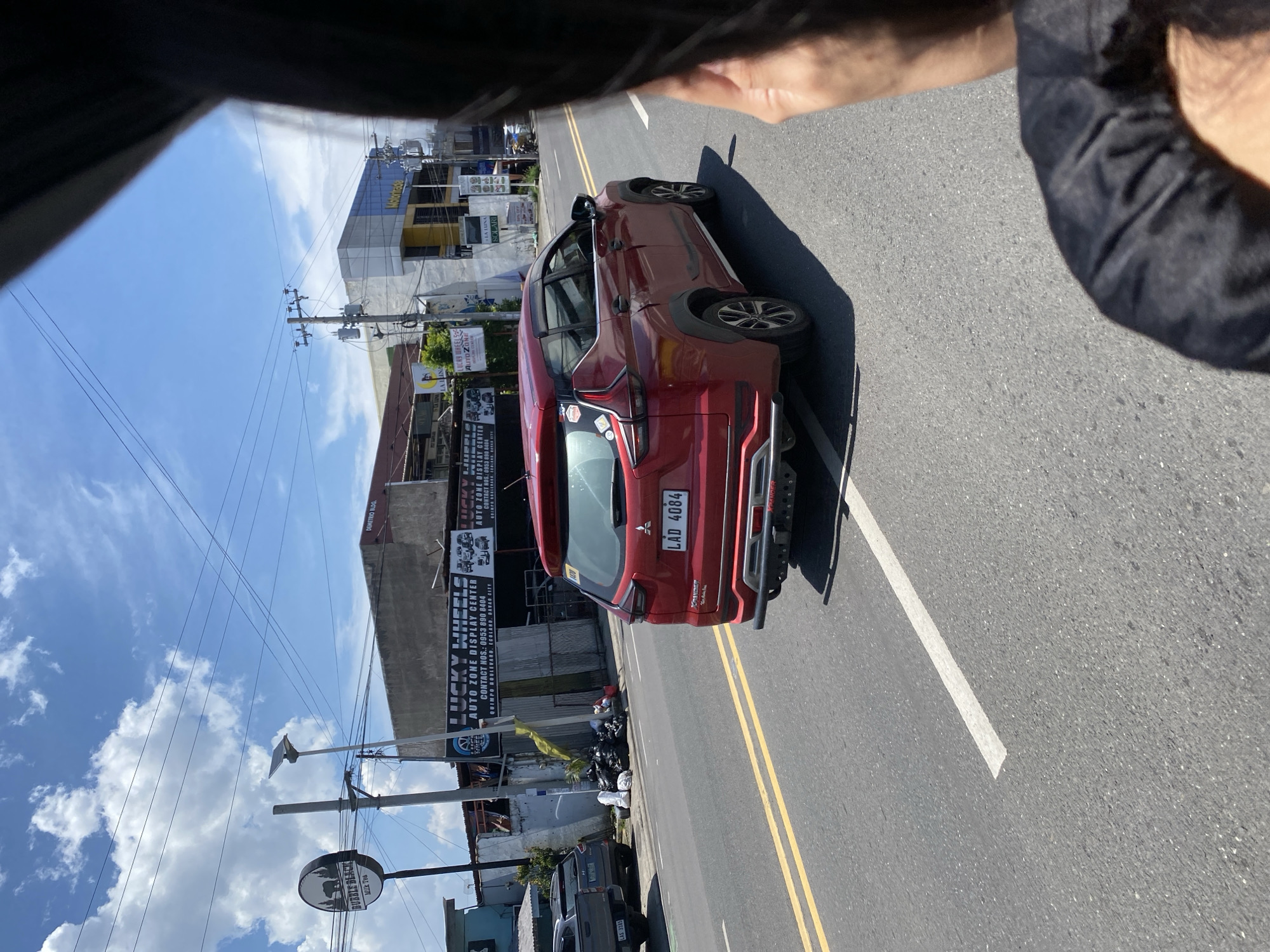 This bright daytime photograph captures an urban street scene in Davao City, Philippines, under a clear blue sky with scattered white clouds. The image is taken from a slightly elevated vantage point, possibly from inside a vehicle, indicated by a dark structure partially obscuring the top left and a visible hand in the top right.
A red SUV, identified by the Philippine license plate "LAD 4084," is parked on the right side of a two-lane asphalt road featuring double yellow center lines and a white shoulder line. Along the sidewalk next to the SUV, piles of black trash bags are visible. Further down the street, a dark pickup truck with license plate "LAS 3185" is partially seen, along with several motorcycles.
The street is lined with buildings housing various commercial establishments. Utility poles with numerous overhead power lines are prominent against the sky. Visible signage includes "LUCKY WHEELS," "AUTO ZONE DISPLAY CENTER," and "DEMETRIO BLDG." A sign also displays "CONTACT NOS.: 0953 890 8404" and the address "QUIMPO BOULEVARD, ECOLAND, DAVAO CITY." In the distance, a blue building bears the name "Motortrada," and a round sign on a pole advertises "Bubble Wrap Milk Tea." Strong sunlight and shadows suggest it is late morning or early afternoon.