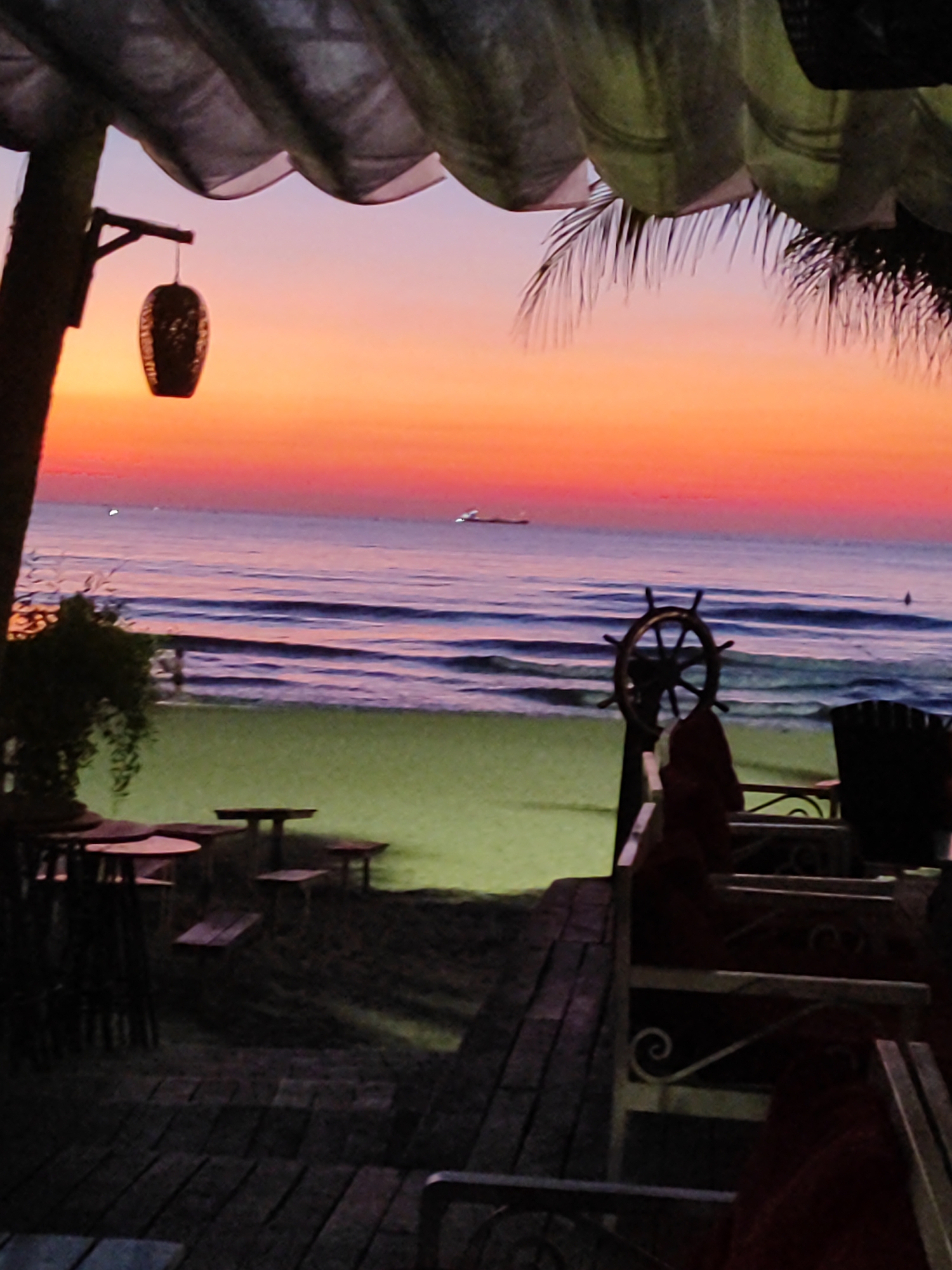 This image depicts a serene sunset scene at a beachside establishment in Da Nang, Vietnam. The foreground features a wooden deck with several tables and chairs, suggesting an outdoor dining or seating area. One of the tables has a rustic, lantern-like decoration hanging from a wooden beam. A ship's wheel is prominently displayed as a decorative element. In the middle ground, a sandy beach with a few scattered tables and chairs transitions into the calm ocean. The water exhibits gentle waves, and in the distance, a large ship is visible on the horizon. The sky is a gradient of warm colors, transitioning from a deep orange and pink near the horizon to a lighter purple and blue overhead, creating a picturesque sunset. The overall atmosphere is peaceful and tranquil, characteristic of a beach setting at dusk.