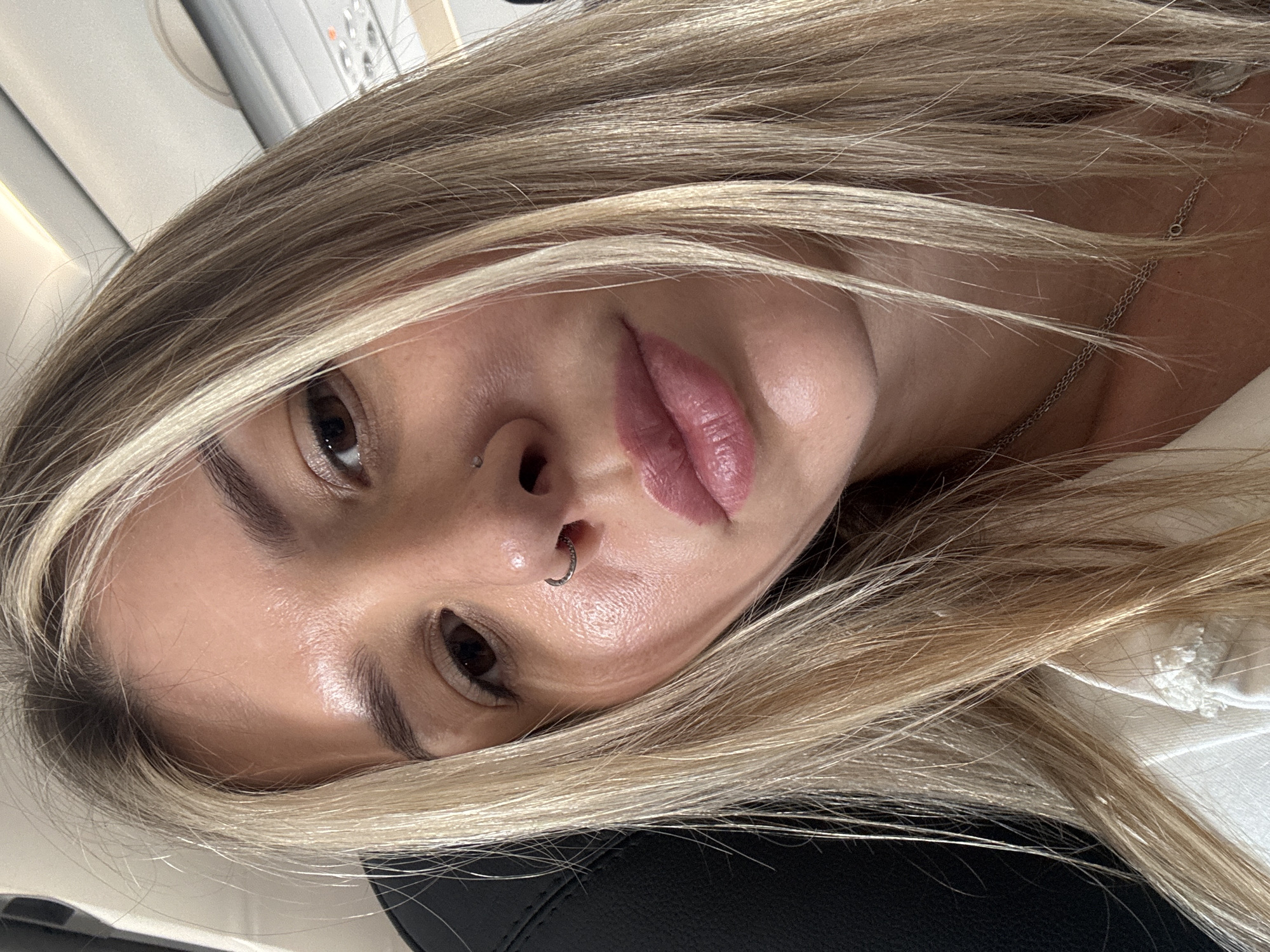 A close-up selfie shows a woman with blonde hair, possibly on an airplane. She is looking at the camera with a neutral expression. Her hair is partially covering her face and shoulders. She is wearing makeup, including lipstick and eye shadow. A nose ring is visible on her left nostril. She is also wearing a silver necklace. The background appears to be the interior of an airplane cabin, with seats and overhead compartments visible.