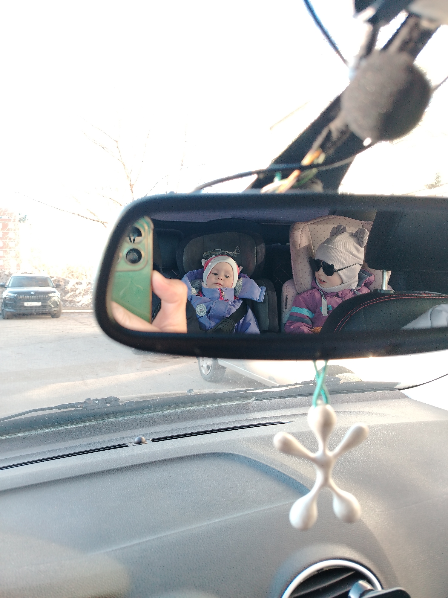 The image, apparently taken from the driver's seat of a car in Chaikovsky, Russia, shows a view through the rearview mirror. Reflected in the mirror are two infants secured in car seats in the back of the vehicle. The infant on the left is wearing a blue snowsuit and hat and appears to be looking at the camera (presumably being held by the person taking the photo). The infant on the right is wearing sunglasses, a pink and purple snowsuit, and a hat with ears. Outside the car, visible through the rearview mirror, is a black car parked on the street. Trees without leaves are also visible in the background. In the foreground, inside the car, is a grey dashboard with an air freshener hanging from the rearview mirror. A smartphone is visible, being used to take the picture. The location is suggested by the presence of cars and street scenes typical of a city like Chaikovsky, Russia.