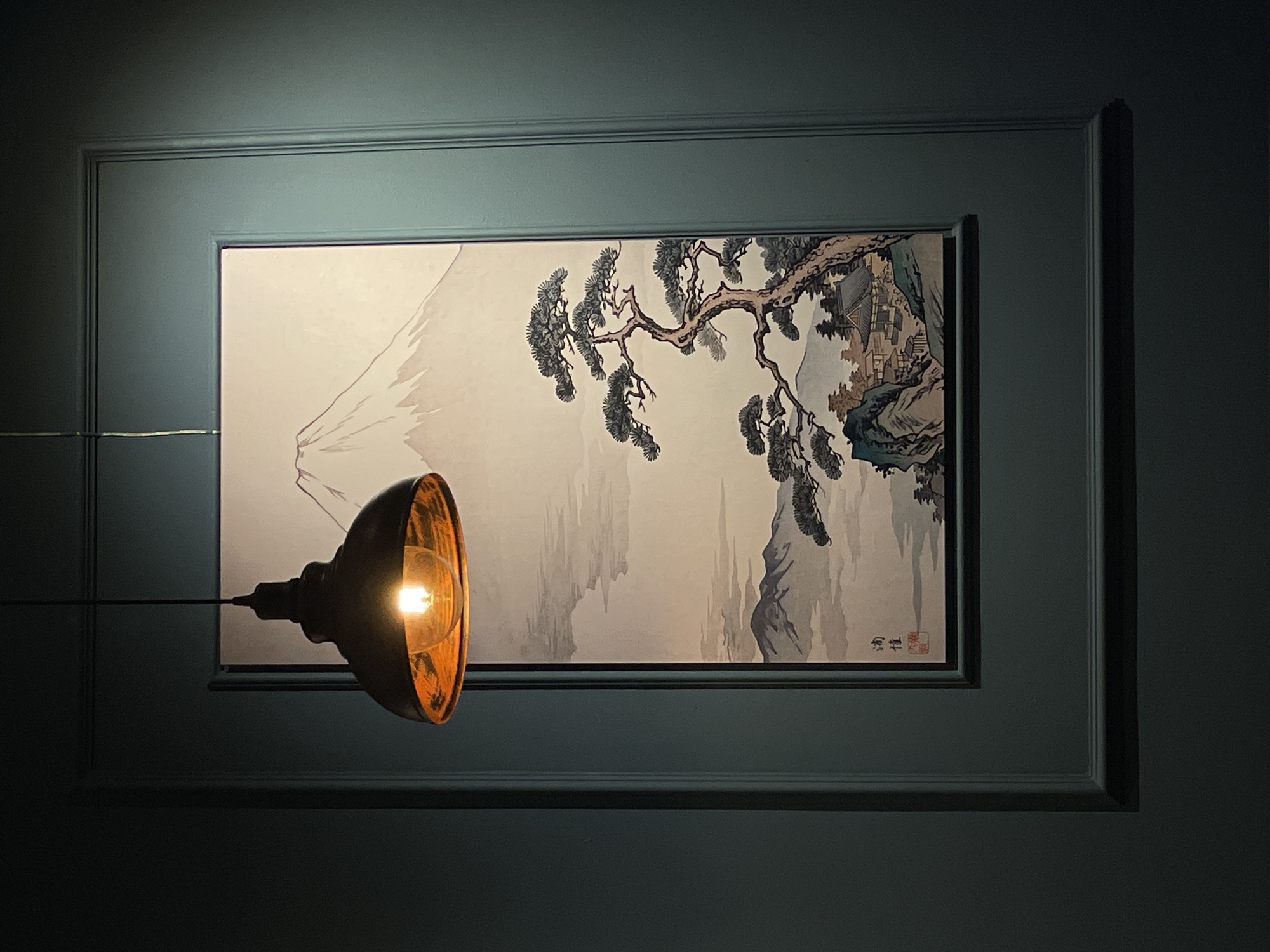 A framed Asian-style landscape painting hangs on a dark, muted teal wall, illuminated by a retro-style light fixture that is mounted to the wall and shines on the artwork. The painting depicts a pale, misty scene with a stylized mountain on the left, possibly meant to evoke Mount Fuji, and a gnarled, aged tree branch with pine needles dominating the right half. Buildings nestle into the landscape under the tree. The light fixture is a dark metal shade, with an exposed glowing filament bulb, and is positioned on the left side of the painting. The scene is dimly lit. The image was taken indoors. There is no evidence in the image that suggests this photograph was taken in Batumi, Georgia.