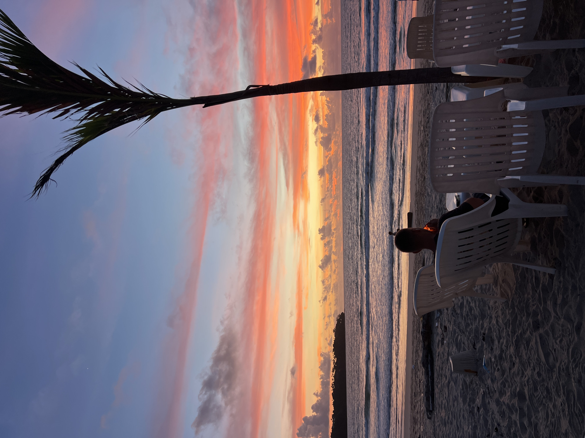 A person is seated in a white plastic chair on a sandy beach, facing the ocean at sunset. The sky is filled with vibrant colors of orange, pink, and blue, with some scattered clouds. A palm tree trunk is visible in the foreground, to the right of the frame. In the distance, a dark, tree-covered landmass is visible on the horizon. Several other white plastic chairs and tables are set up on the beach, suggesting a seating area. The waves are gently rolling onto the shore, reflecting the colorful sky. The overall scene captures a serene and picturesque moment at a beachside location, likely a resort or restaurant in Choeng Thale, Thailand.