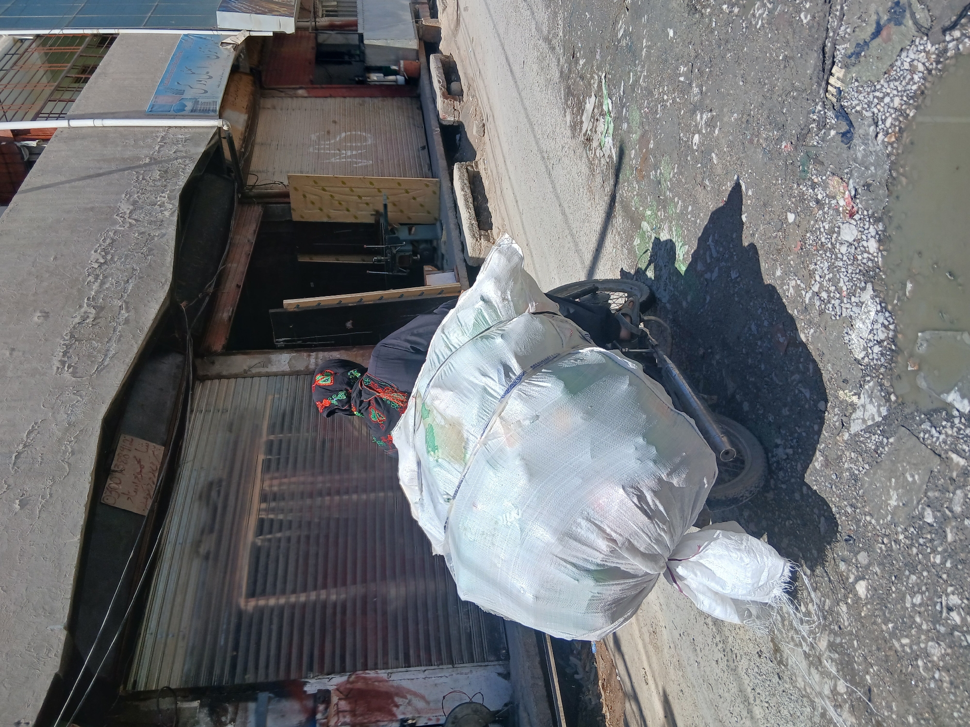 The image captures a person pushing a heavily loaded motorcycle through a narrow, unpaved street or alleyway in Quetta City Tehsil, Pakistan. The scene depicts a bustling, urban environment with concrete buildings and closed shops lining one side.
The central subject is an individual, likely female, wearing a dark garment and a vibrantly embroidered headscarf. They are positioned behind a motorcycle, which is almost entirely obscured by a substantial stack of large white, possibly plastic, bags. These bags appear to be filled with light, bulky materials, likely recyclables or goods being transported for sale or collection. The motorcycle's wheels are visible at the bottom, suggesting it is being pushed or slowly ridden.
The street is rough and uneven, covered with dirt, gravel, and scattered debris. Several muddy puddles are visible, particularly on the right side of the frame, indicating poor drainage or recent moisture. Bright sunlight casts long, distinct shadows of the person and motorcycle on the ground, suggesting it is daytime, possibly morning or late afternoon. The sky is clear, implying sunny weather.
On the left, several buildings feature roll-down metal shutters, indicating closed shops or businesses. Above one of these shops, a blue banner with partially visible text in Urdu/Arabic script can be seen. Another smaller, faded sign on a shop shutter shows the numbers "00 52812" along with some illegible script, potentially a phone number or address. The architecture and street conditions are characteristic of some urban areas in South Asia.