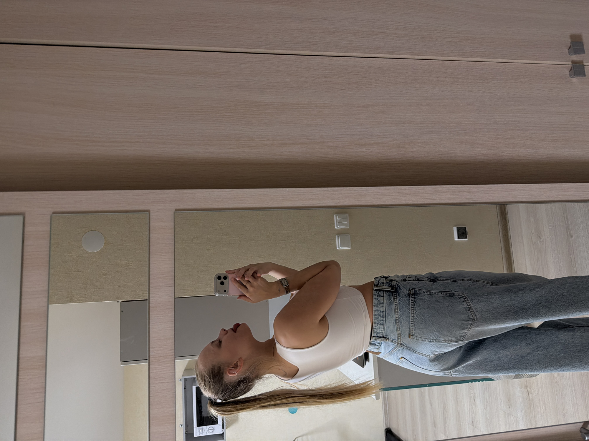 A young woman is taking a selfie in a mirror. She is wearing a white tank top and blue jeans. Her blonde hair is pulled back in a ponytail. The background includes a wardrobe and wall fixtures. The scene appears to be indoors, possibly in a residential setting. The location in Izhevsk, Russia is not identifiable from the image alone.