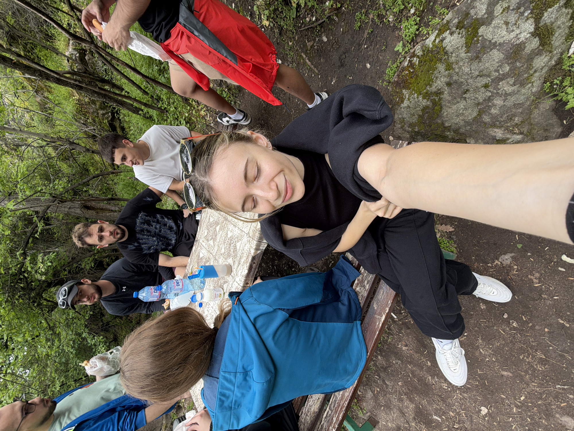 A group of people are gathered around a table outdoors in Ustina, Bulgaria. One woman with blonde hair is taking a selfie while sitting on a bench. She is wearing sunglasses, a black shirt, black pants, and white sneakers. Several other people are seated or standing around the table, which has water bottles and other items on it. The surrounding environment includes trees, foliage, and a dirt ground.