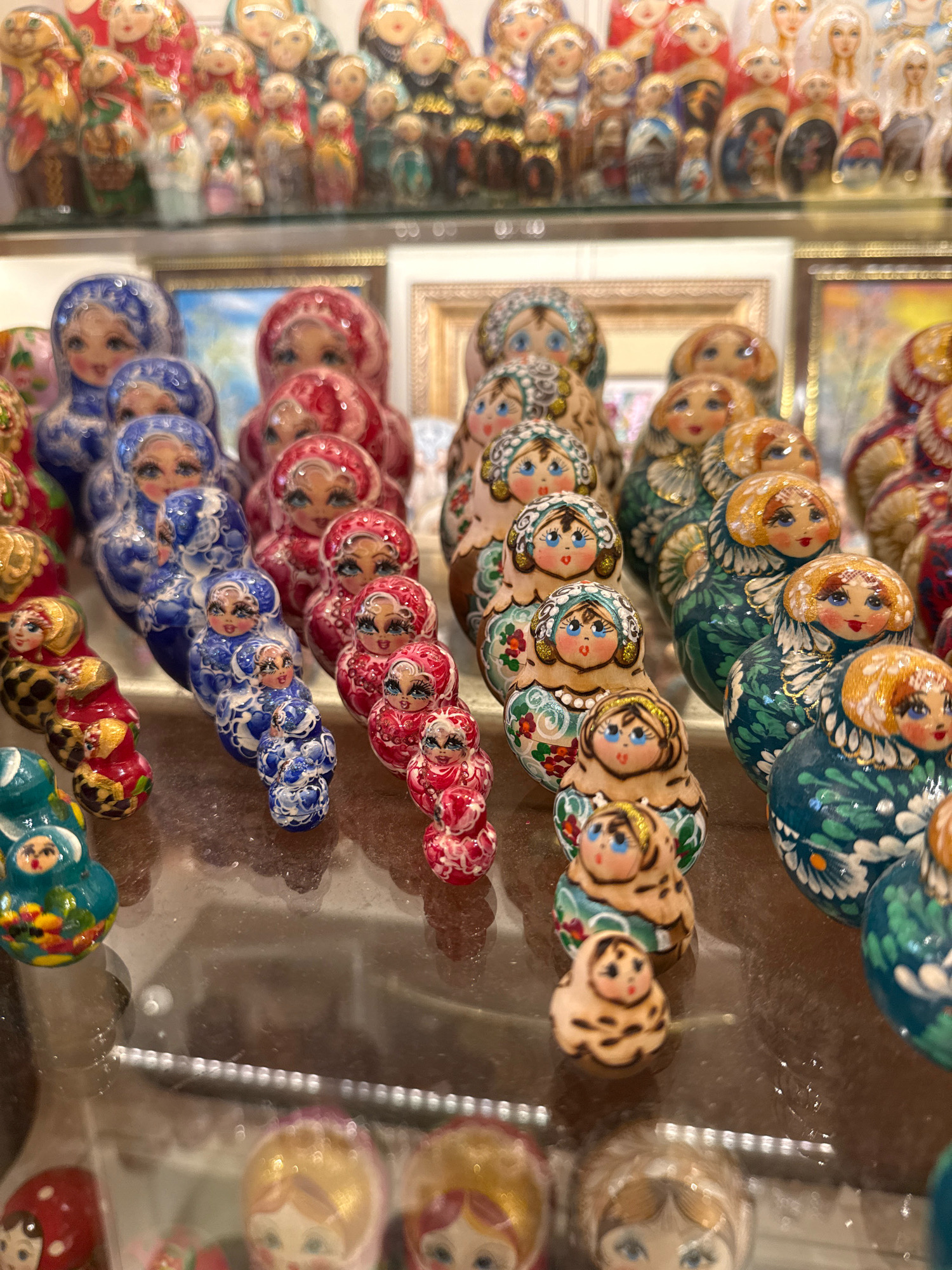 A collection of nesting dolls, known as Matryoshka dolls, is displayed on a multi-tiered shelf in a brightly lit store. The dolls are arranged in rows, with each doll featuring a painted face and traditional Russian attire. The dolls vary in size and color, with some in shades of red, blue, and green, and others with intricate patterns. The background reveals other displays of similar dolls and framed artwork, suggesting a souvenir or craft shop. The lighting is bright and even, illuminating the details of the dolls and their surroundings. The overall scene is one of commercial display and cultural representation, showcasing the artistic heritage of Russia.