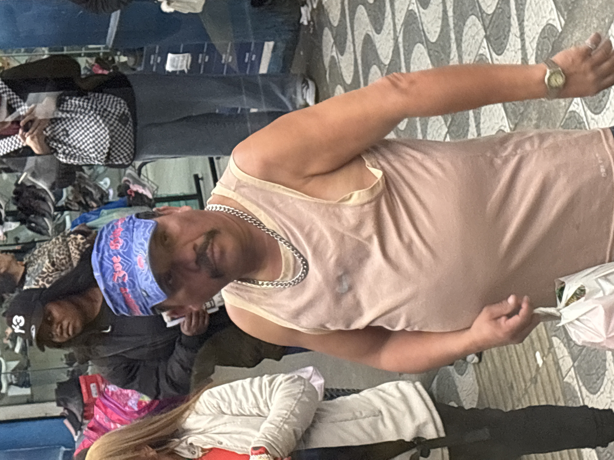 In São Paulo, Brazil, a man wearing a light pink tank top and a blue bandana stands on a sidewalk with a patterned tile design. He has a mustache and is wearing a silver chain necklace and a wristwatch. He is holding a white plastic bag in his right hand. To his left, a woman in a light-colored jacket is partially visible. In the background, other individuals are present, along with displays of shoes and bags, suggesting a commercial area, possibly a street market or a shop. The lighting suggests it is daytime.