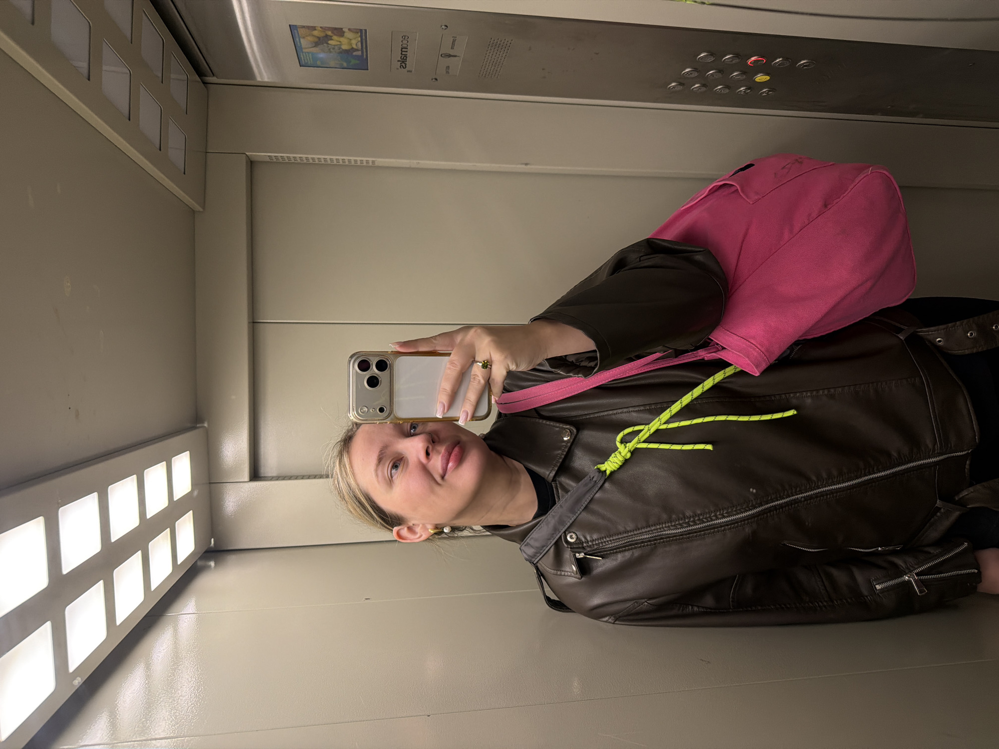 A fair-skinned woman is taking a selfie in an elevator. She is wearing a brown leather jacket with a bright yellow drawstring, and a pink backpack is slung over her shoulder. She is holding a smartphone in her left hand, visible in the selfie. The interior of the elevator has lights, buttons, and signage. The elevator is located in Chaikovsky, Russia.