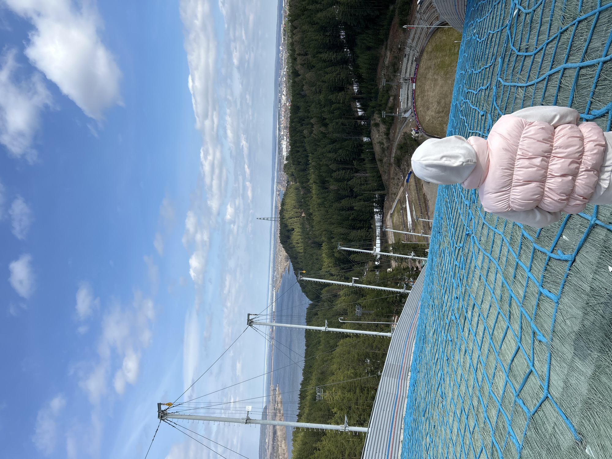 A low-angle, eye-level shot captures a child wearing a pink puffy jacket and white hat, positioned against a blue net structure, with a broad cityscape in the background. The child is viewed from the back, with their head and shoulders visible.
The backdrop reveals a dense forest transitioning into an urban area, characterized by buildings and infrastructure. In the distance, a body of water stretches towards the horizon under a partly cloudy sky. The sky is bright blue, punctuated with cumulus clouds.
The overall perspective indicates a high vantage point, suggesting the photo was taken from an elevated structure. This could be a ski jump ramp, given the net structure's appearance, with the city of Прикамский, Russia, providing the geographical context for the image.