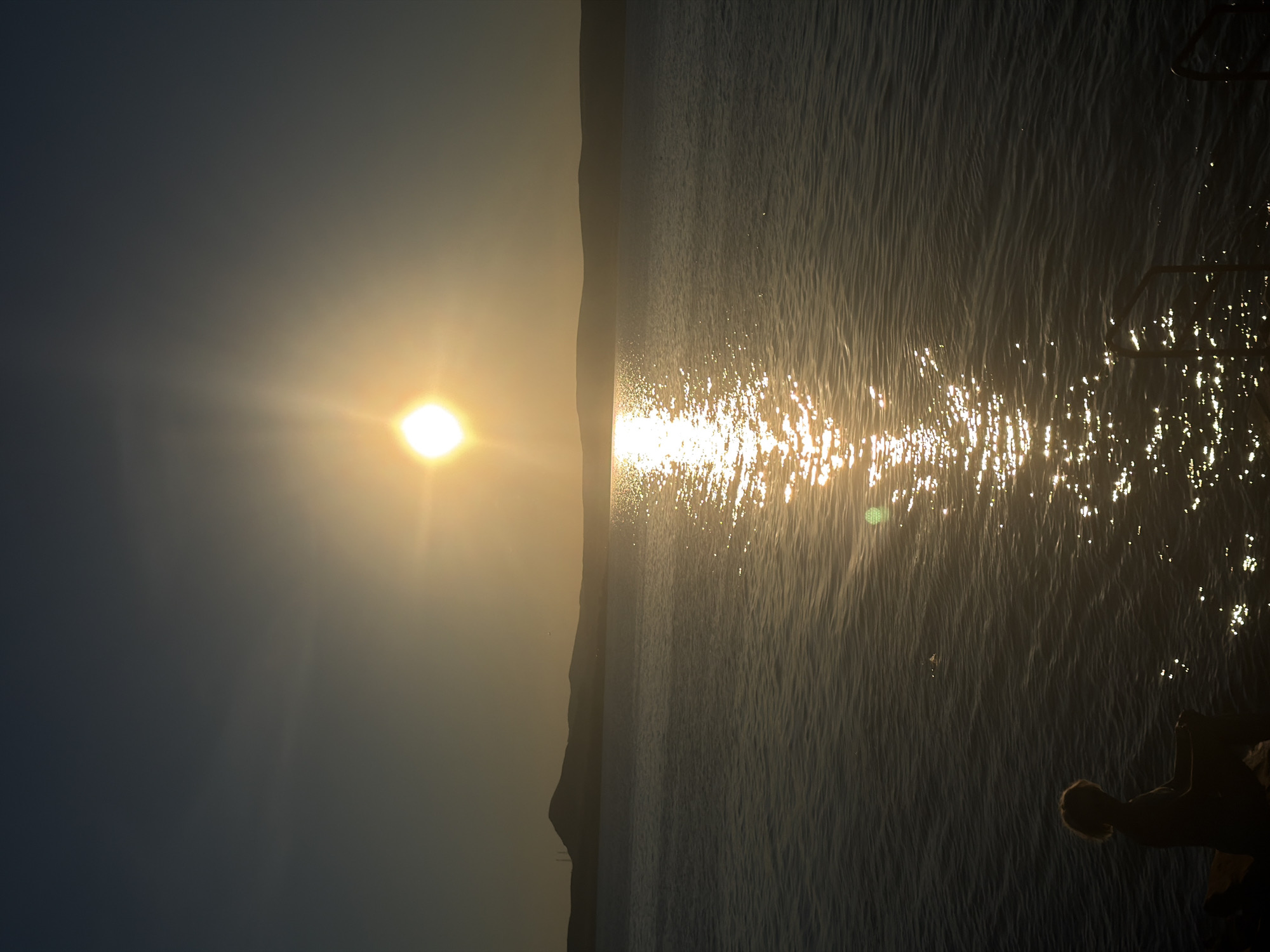 A wide shot captures a tranquil sunset over a body of water, possibly a lake or sea, with a distant, silhouetted hilly landscape. The sun, a bright orb radiating light, is positioned high in the sky, casting a shimmering golden path across the water's surface. In the foreground, to the left, a lone figure, silhouetted by the setting sun, sits on the shore, their back to the camera. Their hair is light and appears to be tied up, and they are dressed in dark clothing. The water is calm, with gentle ripples reflecting the sun's glow. To the right of the frame, a metal railing, likely part of a pier or dock, emerges from the water. The sky transitions from a clear, pale blue at the top to a warmer, golden hue near the horizon. The overall atmosphere is peaceful and serene. The image's location is described as unknown, Russia.