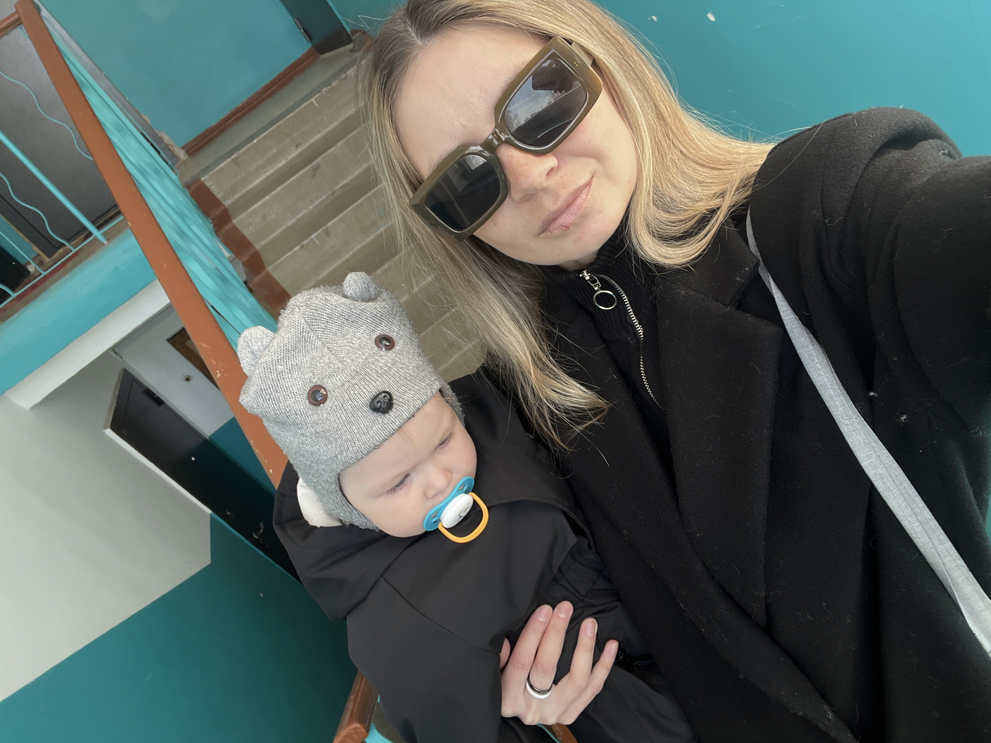 A selfie-style portrait shows an adult female holding an infant in an indoor setting within Chaikovsky, Russia. The female, positioned in the foreground, has blonde hair, wears brown sunglasses, a black zippered top, and a black coat. A black and white ring is visible on her left ring finger. She holds an infant dressed in black outerwear and a grey knitted hat with bear ears and button eyes. The infant is asleep, resting against the female's chest, with a blue and orange pacifier in its mouth. The background consists of a stairwell, characterized by bright teal-painted walls, a portion of a white ceiling or upper wall, a brown wooden handrail, and visible concrete steps.