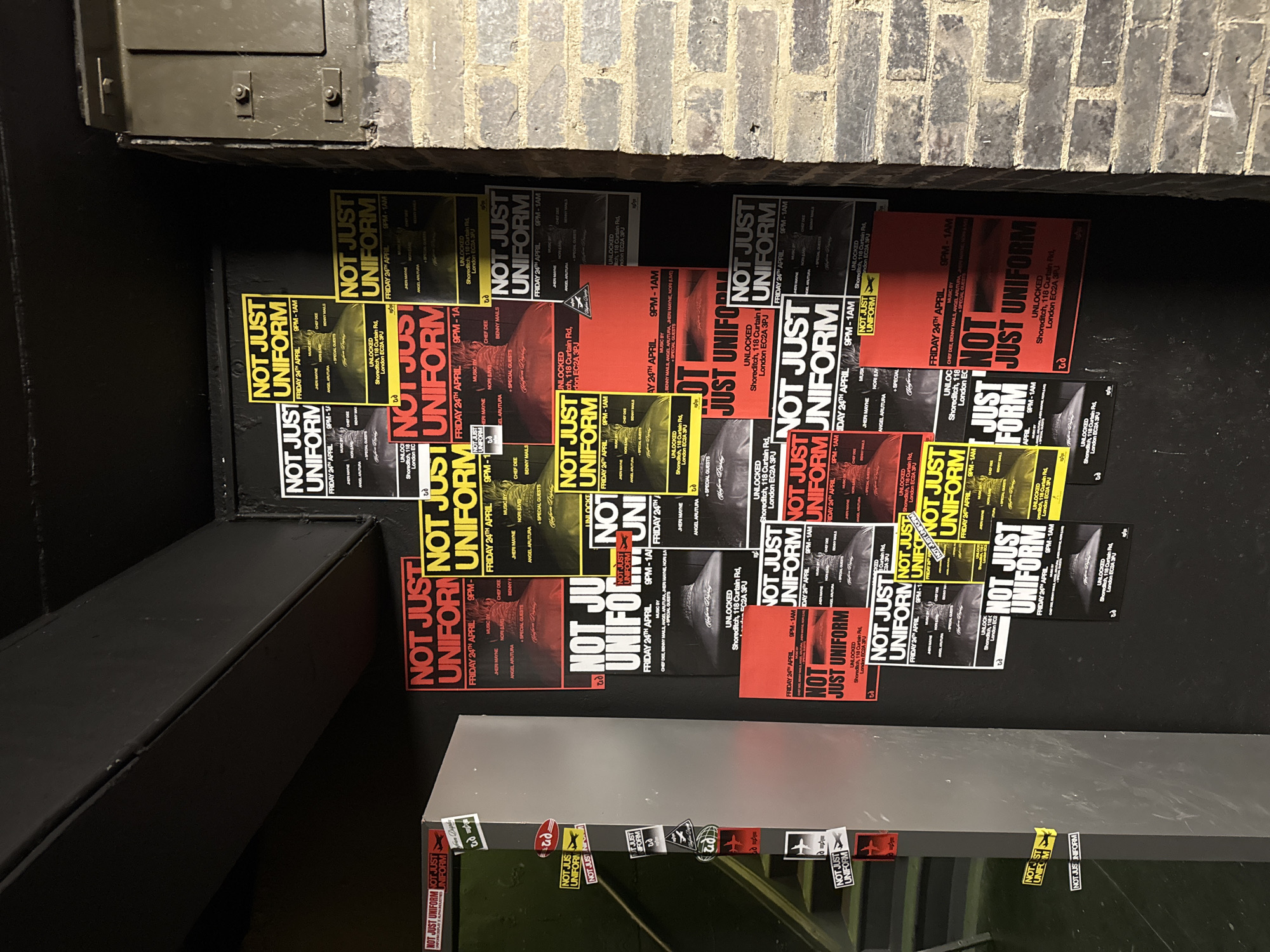 The image captures an outdoor wall, likely in an urban alleyway or building exterior, heavily covered with event posters and stickers. The setting appears dark, with hints of a brick wall in the upper right and a dark metallic structure to the left. A reflective surface is visible at the bottom of the frame, also adorned with stickers.
The primary objects are numerous copies of event posters advertising an event titled "NOT JUST UNIFORM." These posters are displayed in a collage style, overlapping each other, creating a dense and textured surface. There are two main color schemes for the posters: some are predominantly red with black and white text, while others feature a yellow/green gradient with black and white text.
The visible text, repeated across many posters, details the event:
- **Event Name:** NOT JUST UNIFORM
- **Date:** FRIDAY 24TH APRIL
- **Time:** 9PM - 1AM
- **Venue:** UNLOCKED, Shoreditch, 118 Curtain Rd, London EC2A 3PJ
- **Music By:** CHEF DEE, BENNY MAILS, JHERI MAYNE, ANGEL ARUTURA, NORI (LIVE), and SPECIAL GUESTS.
The posters also feature a small graphic, possibly a stylized paper airplane. Various small stickers are also present on the reflective surface below the main poster display, many featuring the "NOT JUST UNIFORM" logo or a similar plane icon. The scene suggests an active urban environment where events are promoted through street art and poster campaigns. No people are visible.