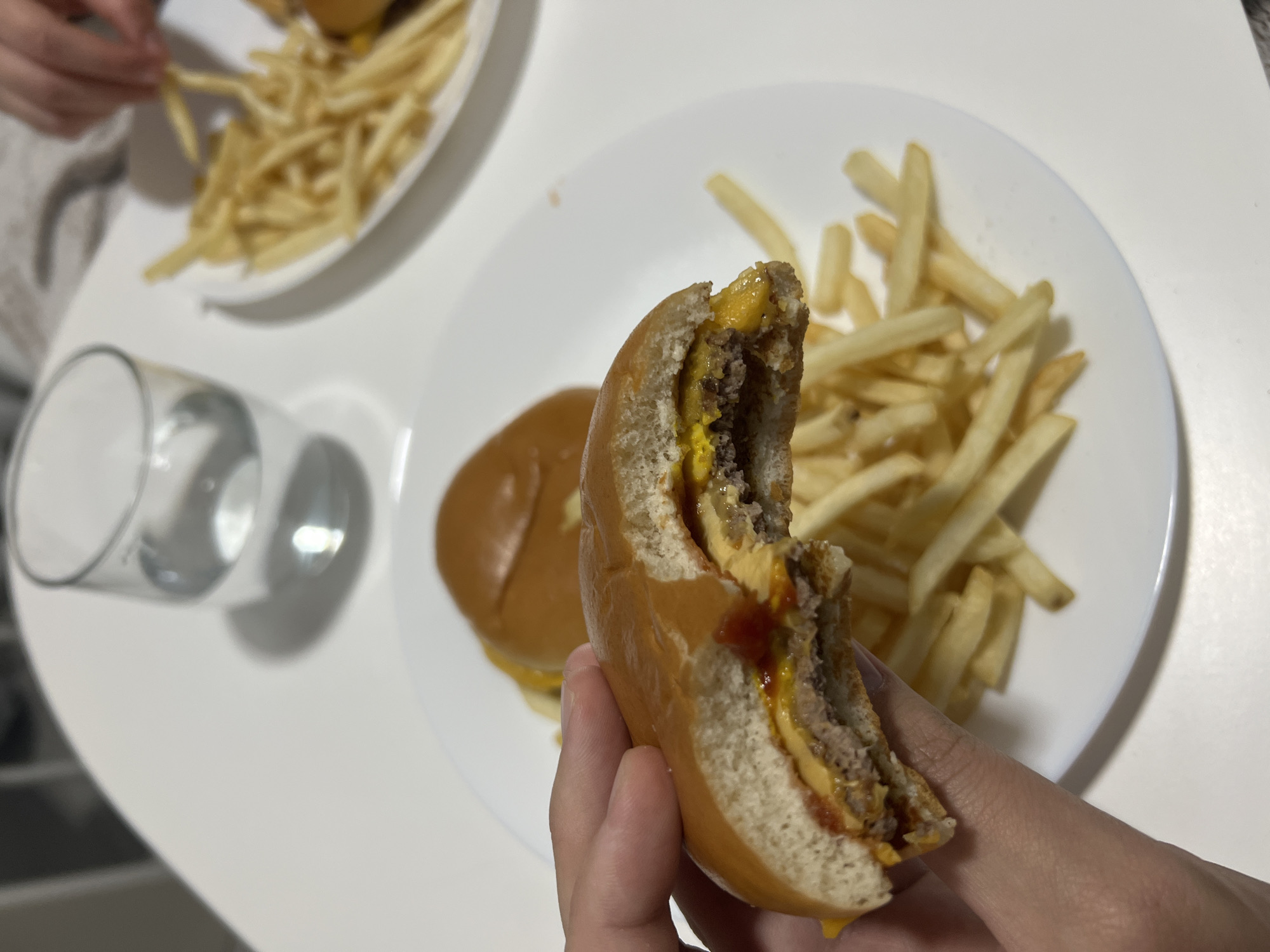 A first-person perspective shot shows a partly eaten double cheeseburger held in a hand. The burger shows a patty with melted cheese, mustard, and ketchup between a sliced bun. The bite reveals the interior of the burger. The burger is positioned over a white plate containing french fries and a second burger. The background shows a clear drinking glass and another plate with french fries. The setting is a white table. The location and date are not discernible. The image contains a person, burgers, fries, a plate, and a glass. It is a meal. Mogán, Spain, cannot be determined from this photo alone.